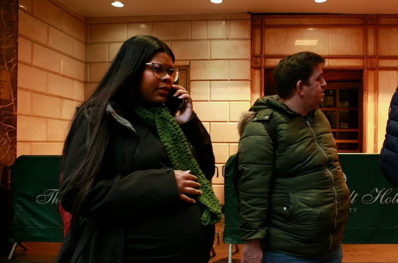 A pregnant woman with glasses and long black hair wearing a green scarf over black clothes talks on a cell phone.