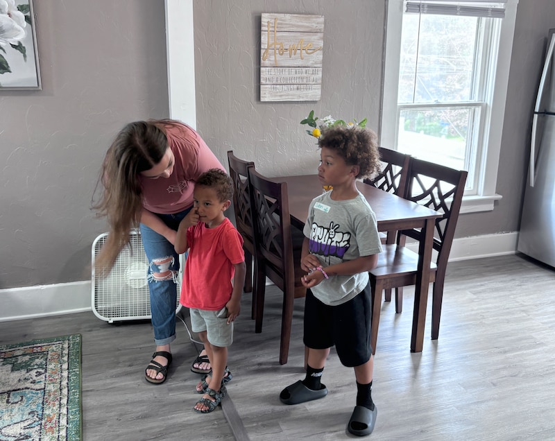 A photograph of a woman with two young children standing inside of a house.