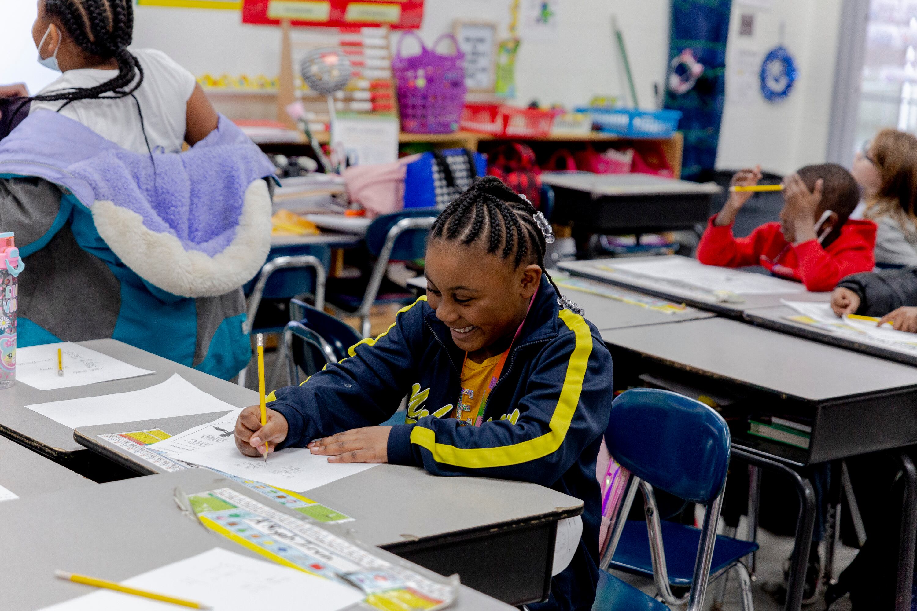 A student with cornrows and a sports jacket smiles as she sits at a desk and writes on a piece of paper