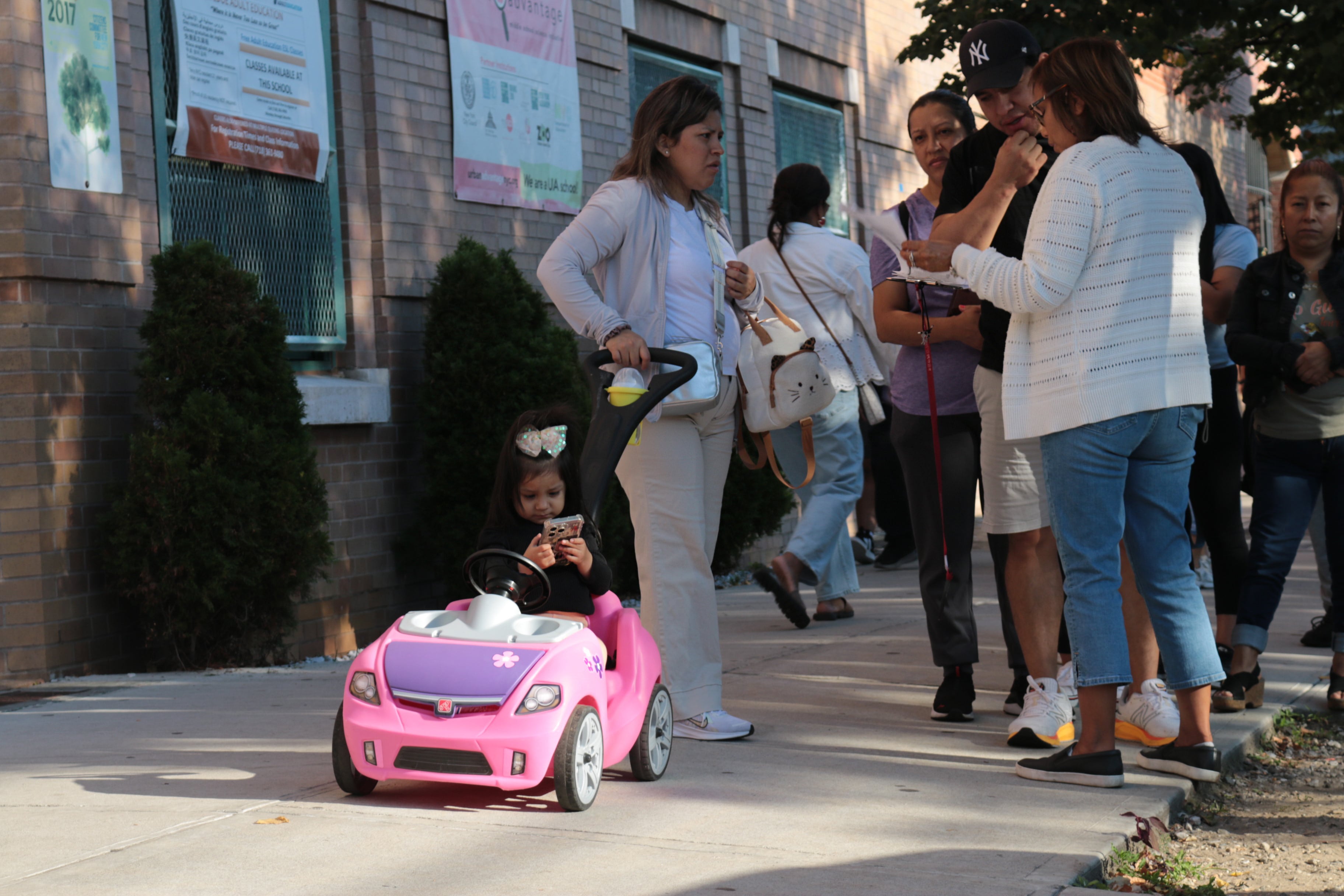 A photograph of a young child looking at a cellphone while sitting in a small toy car while a group of adults stand nearby.