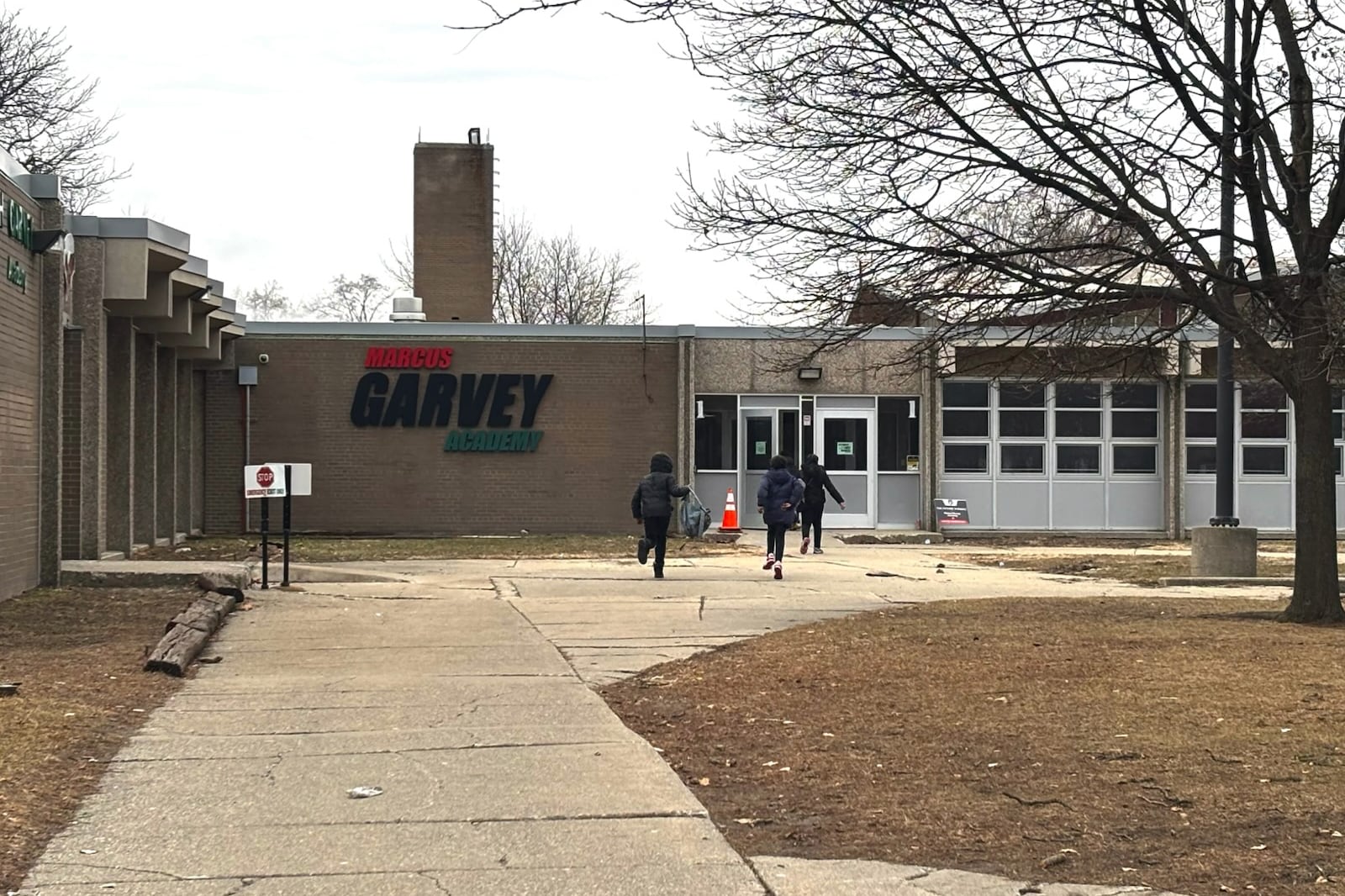 A photograph of three students running toward the front entrance of a school building. All three students are wearing winter coats on a cold and cloudy day.