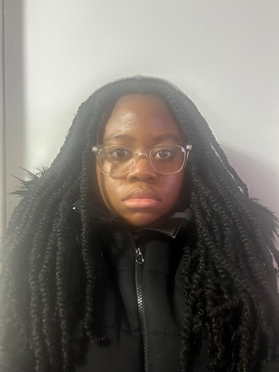 A photograph of a Black high school junior with long dark braids wearing a dark sweater looks at the camera while posing for a portrait.