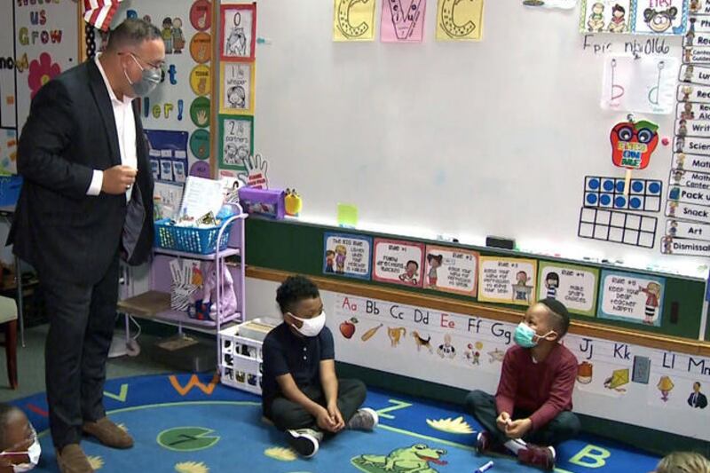Secretary of Education Miguel Cardona, wearing a mask and standing, speaks in a classroom with elementary students, who are sitting on a rug and looking up at him.