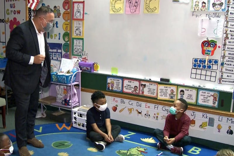 Secretary of Education Miguel Cardona, wearing a mask and standing, speaks in a classroom with elementary students, who are sitting on a rug and looking up at him.