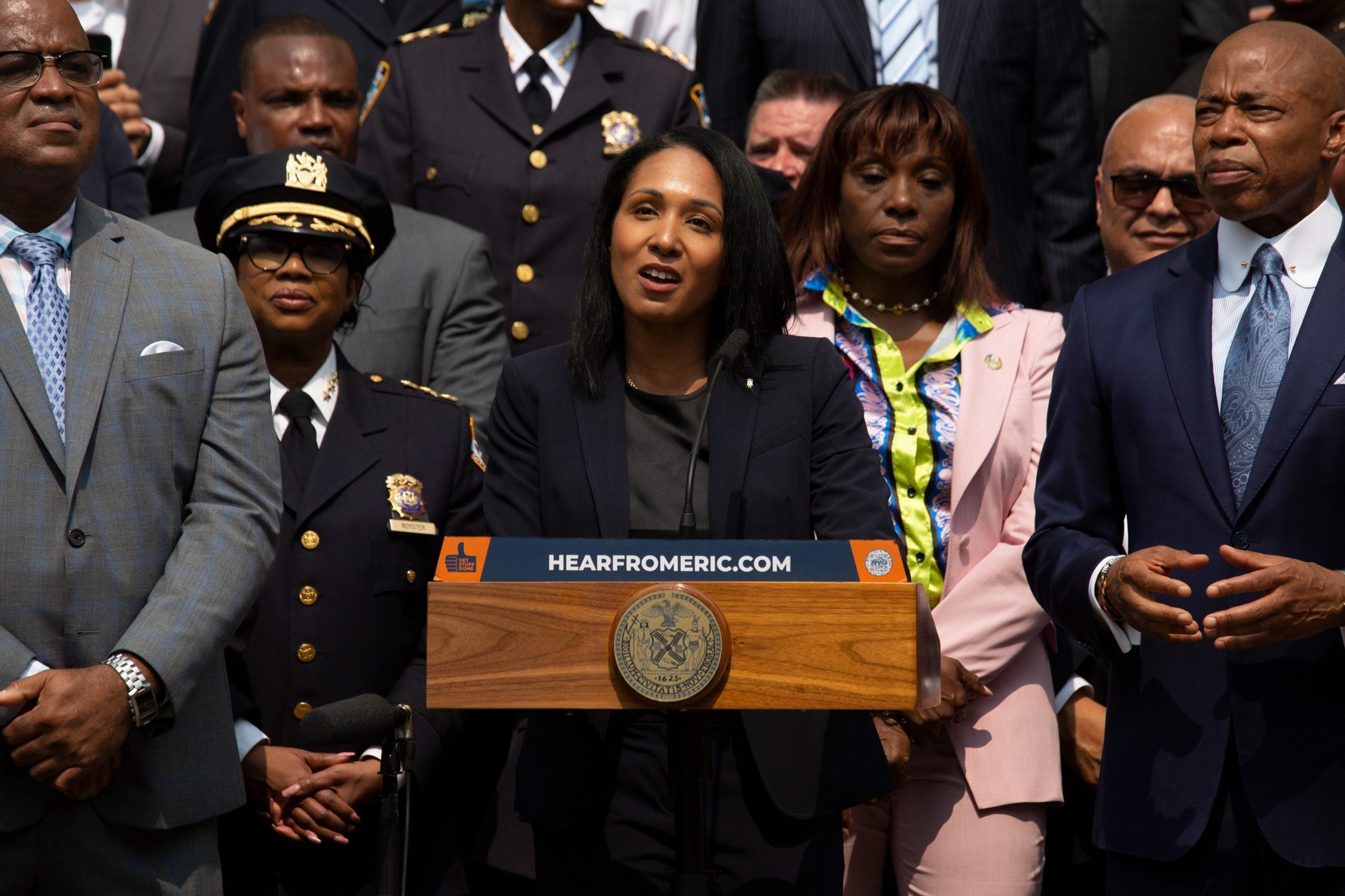 A photograph of a group of adults in suits standing behind a Black woman speaking from a podium.