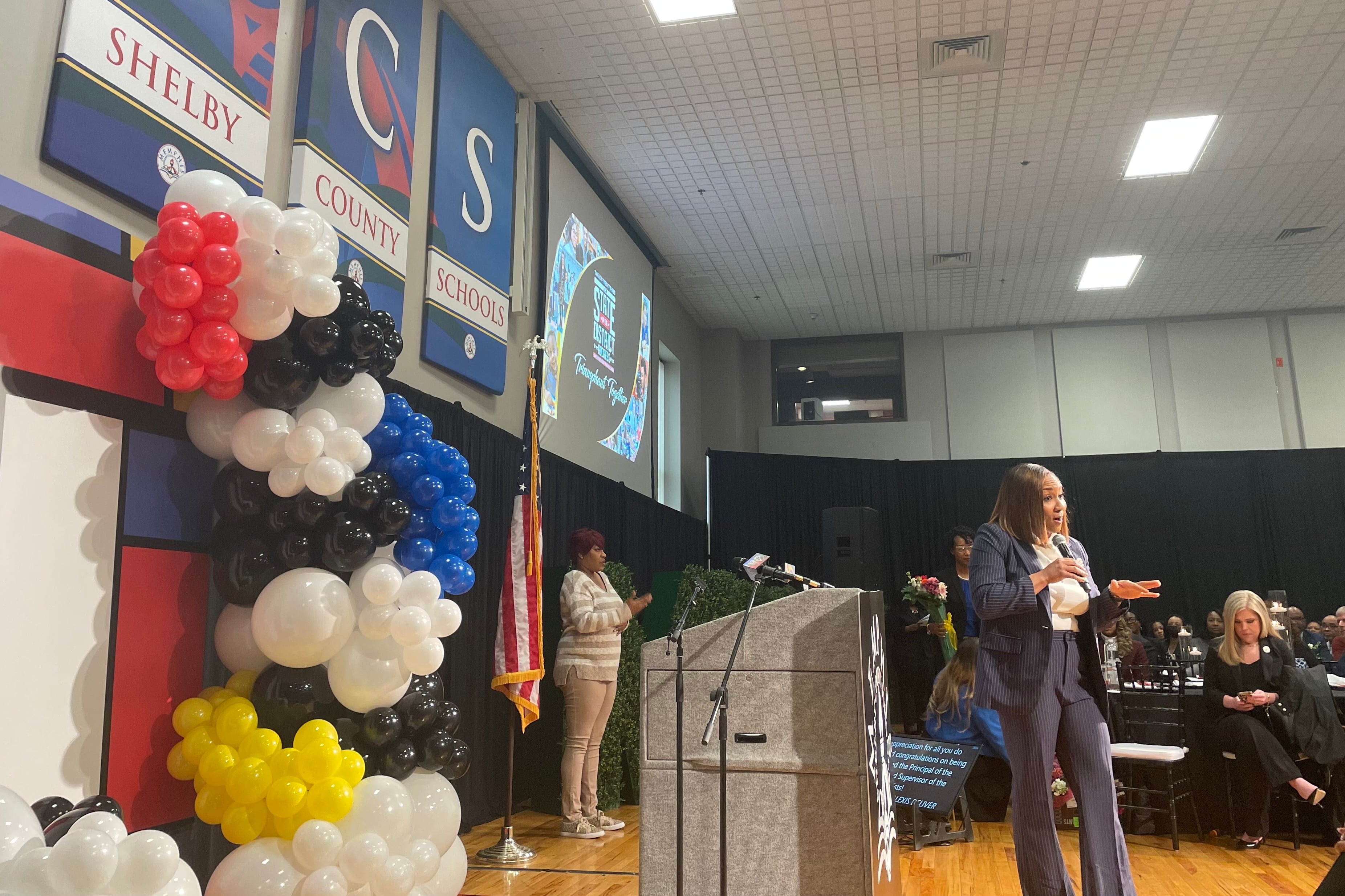 A woman in a gray suit holds a microphone and stands on a school auditorium stage.