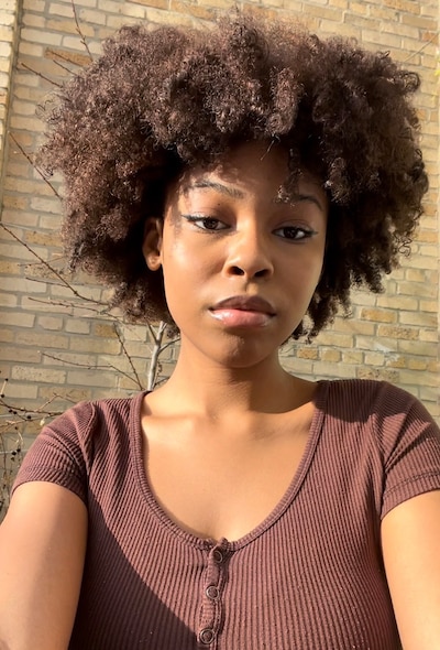 A high school student with brown hair and a brown shirt poses for a portrait with tan bricks in the background.