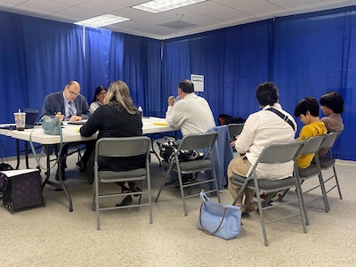 Four people sit across from each other at a table while three people sit in folding chairs in a row in front of the table and a blue curtain in the background.