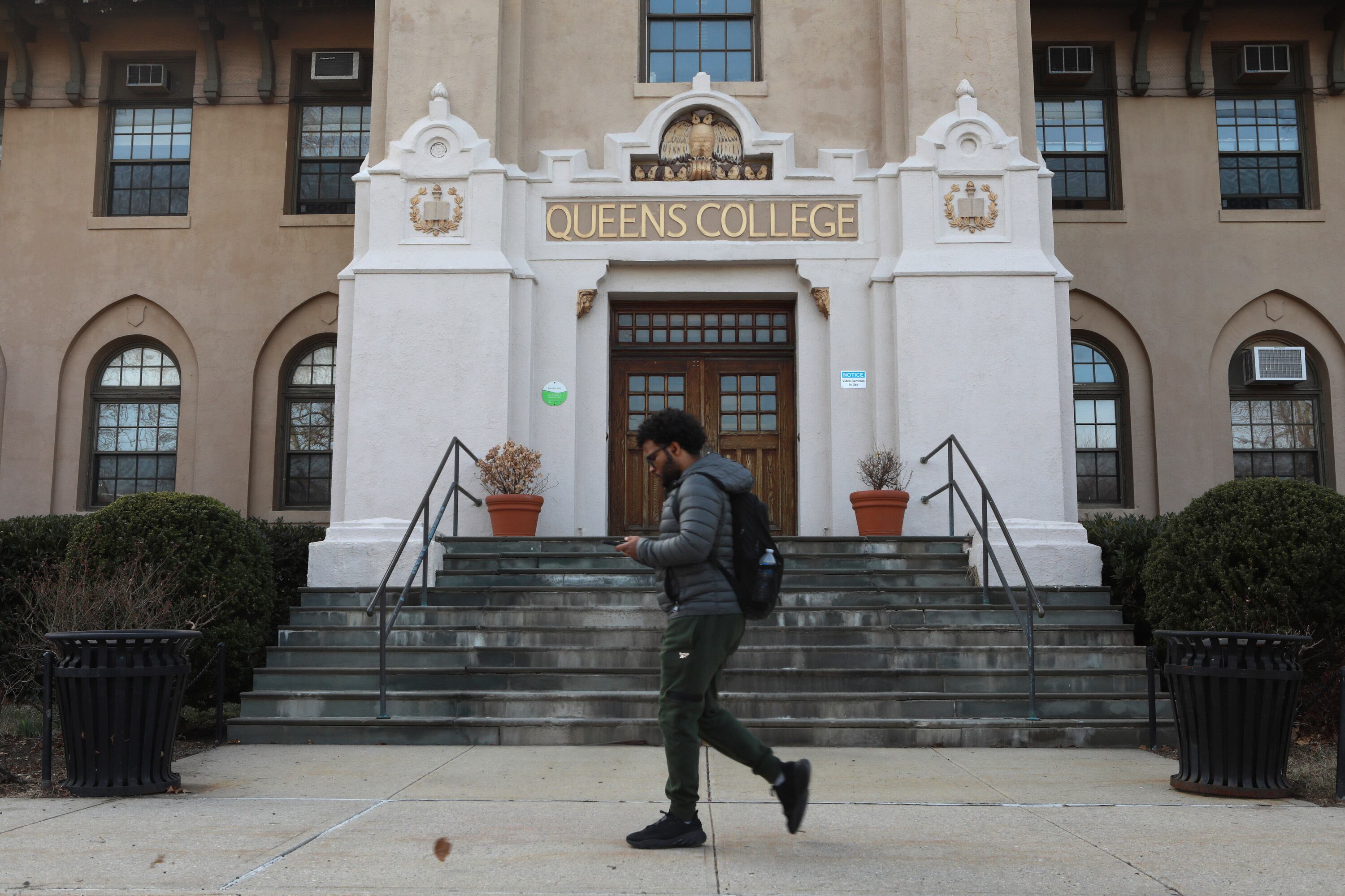A student with short hair and wearing a backpack walks in front of an entrance of a college.