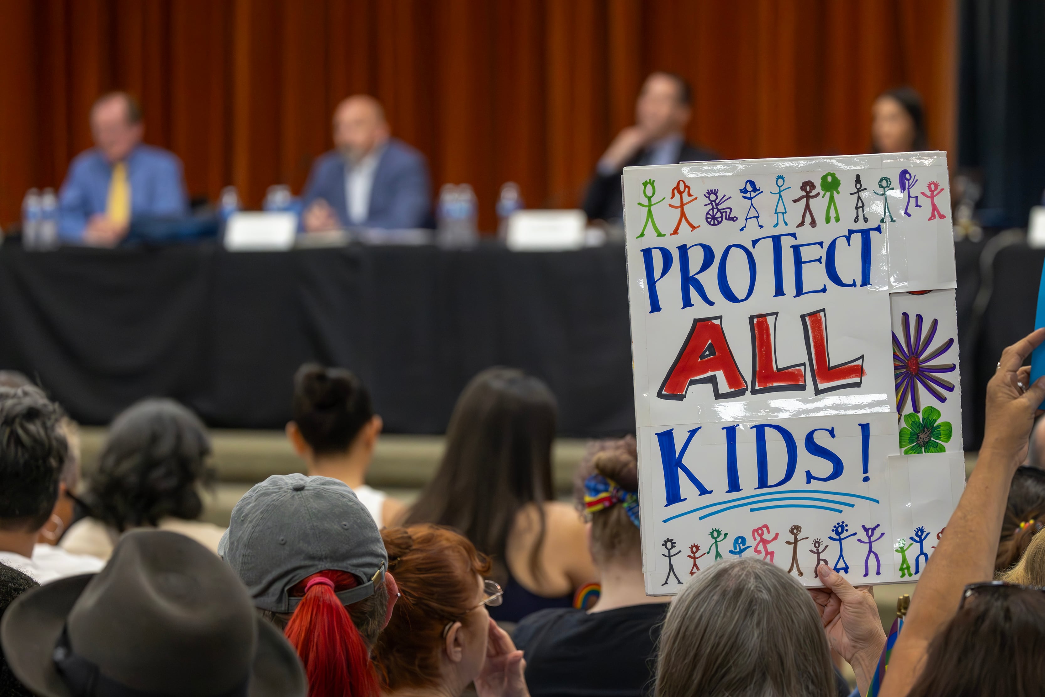 A sign that reads "Protect All Kids!" pops up from a crowd of people sitting in front of a group of people wearing suits and sitting on a stage.