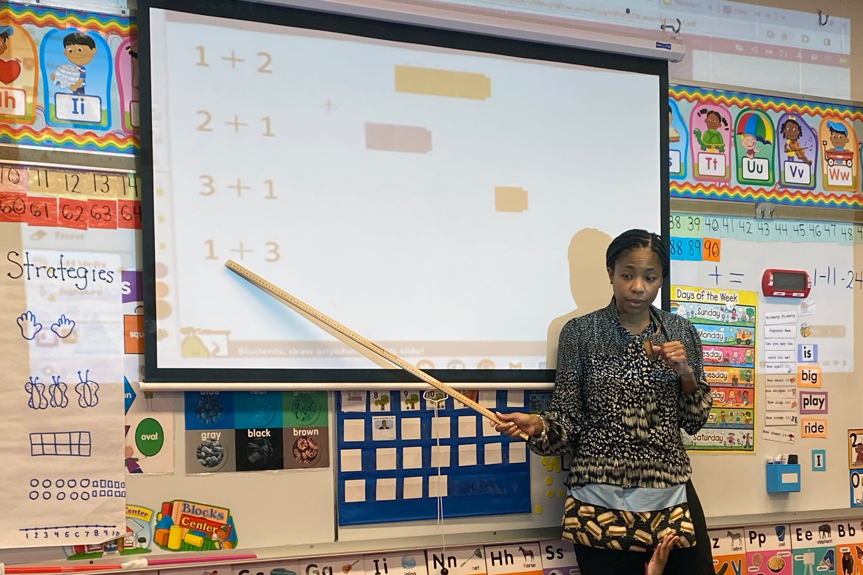 A teacher with dark hair and holding a meter ruler, points to a projector screen on the wall while a group of young students sit on the floor.