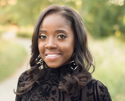 A woman with medium length dark hair and wearing a black blouse poses for a photo with a large smile in front of a green background.