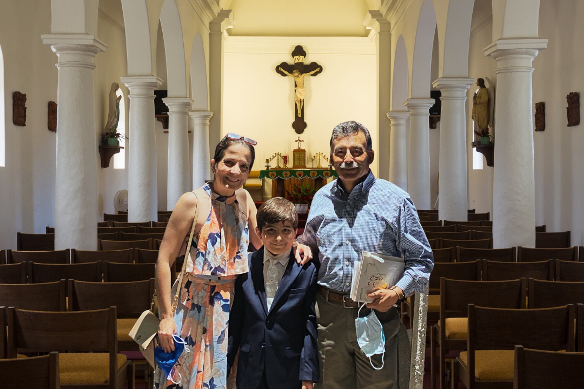 A mother and father stand in a church with a 10-year-old boy between them. They all smile at the camera. A crucifix can be seen in the background.