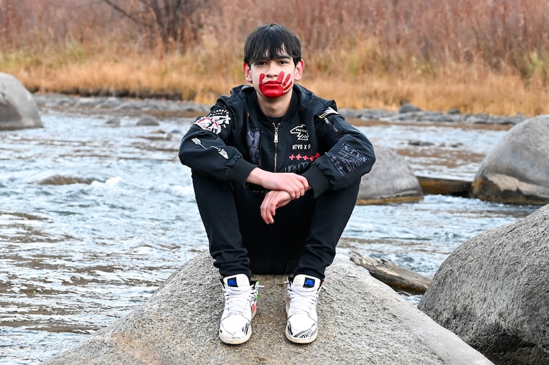 A boy wearing a black jacket and pants, white shoes and has a red handprint across his face sits on a rock with a stream in the background while posing for a photograph.