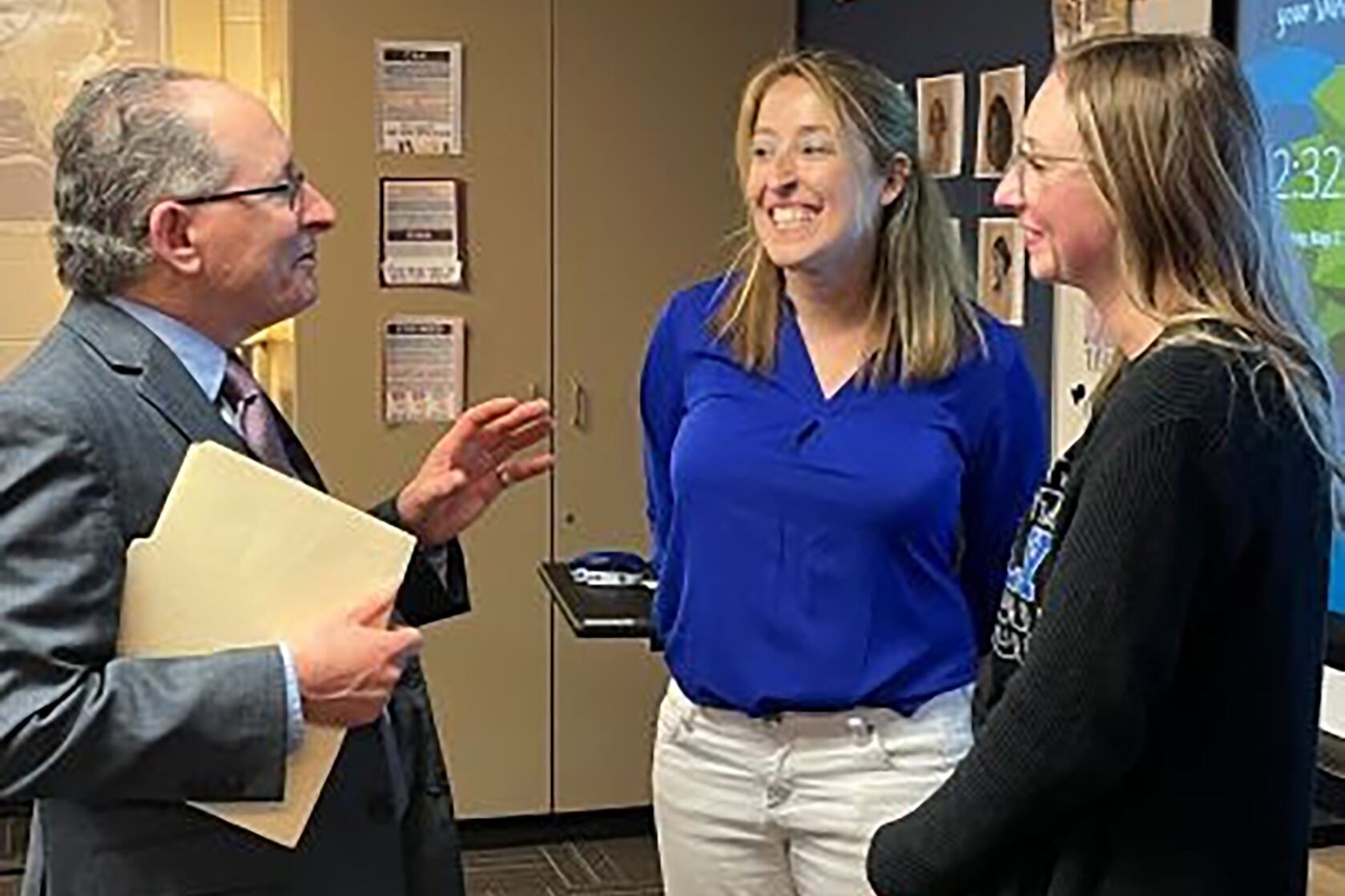 A man in a gray suit and glasses talks to a woman with a blue top and white pants and a woman with a black top.