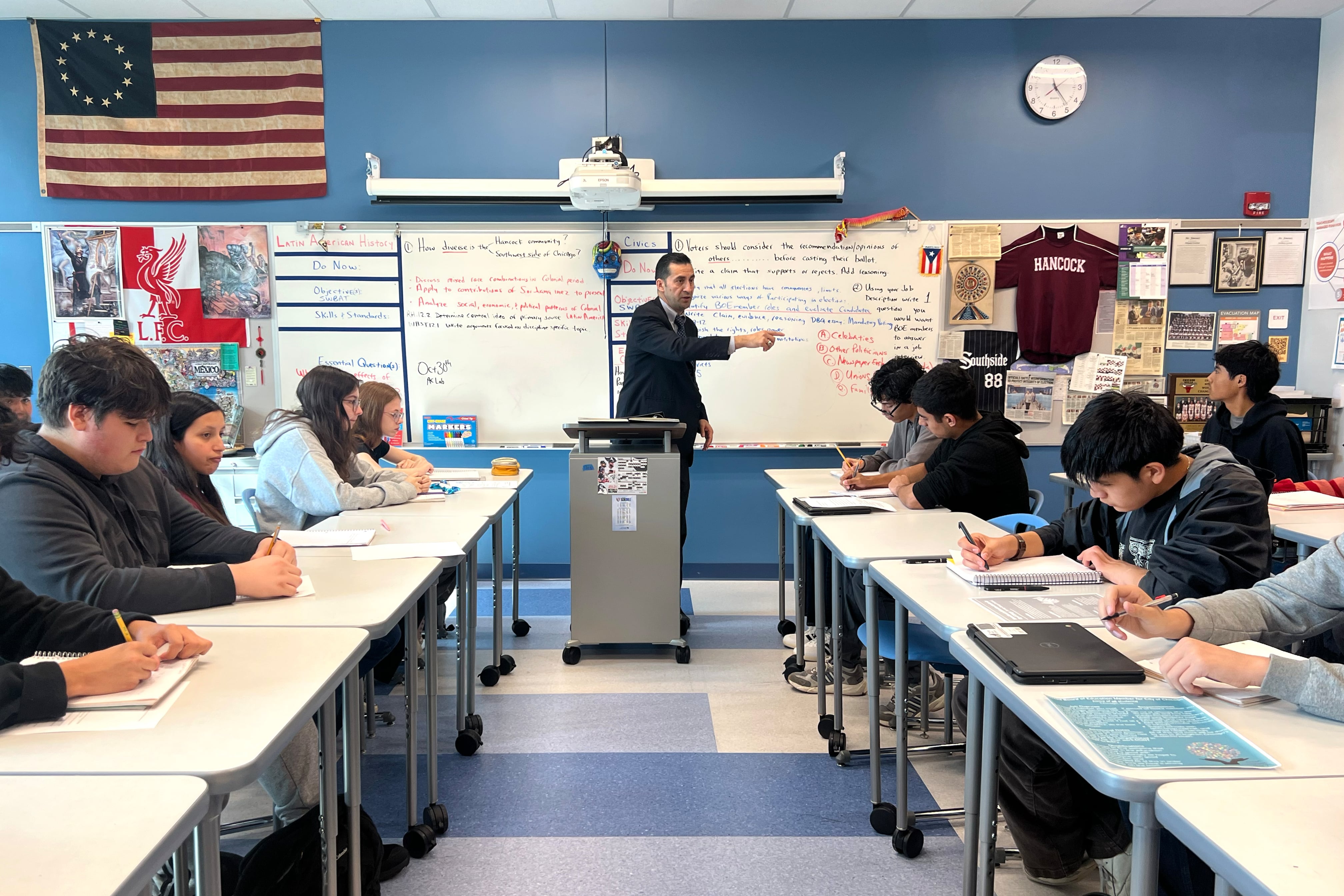 A man in a suit jacket and tie points in a classroom of students.