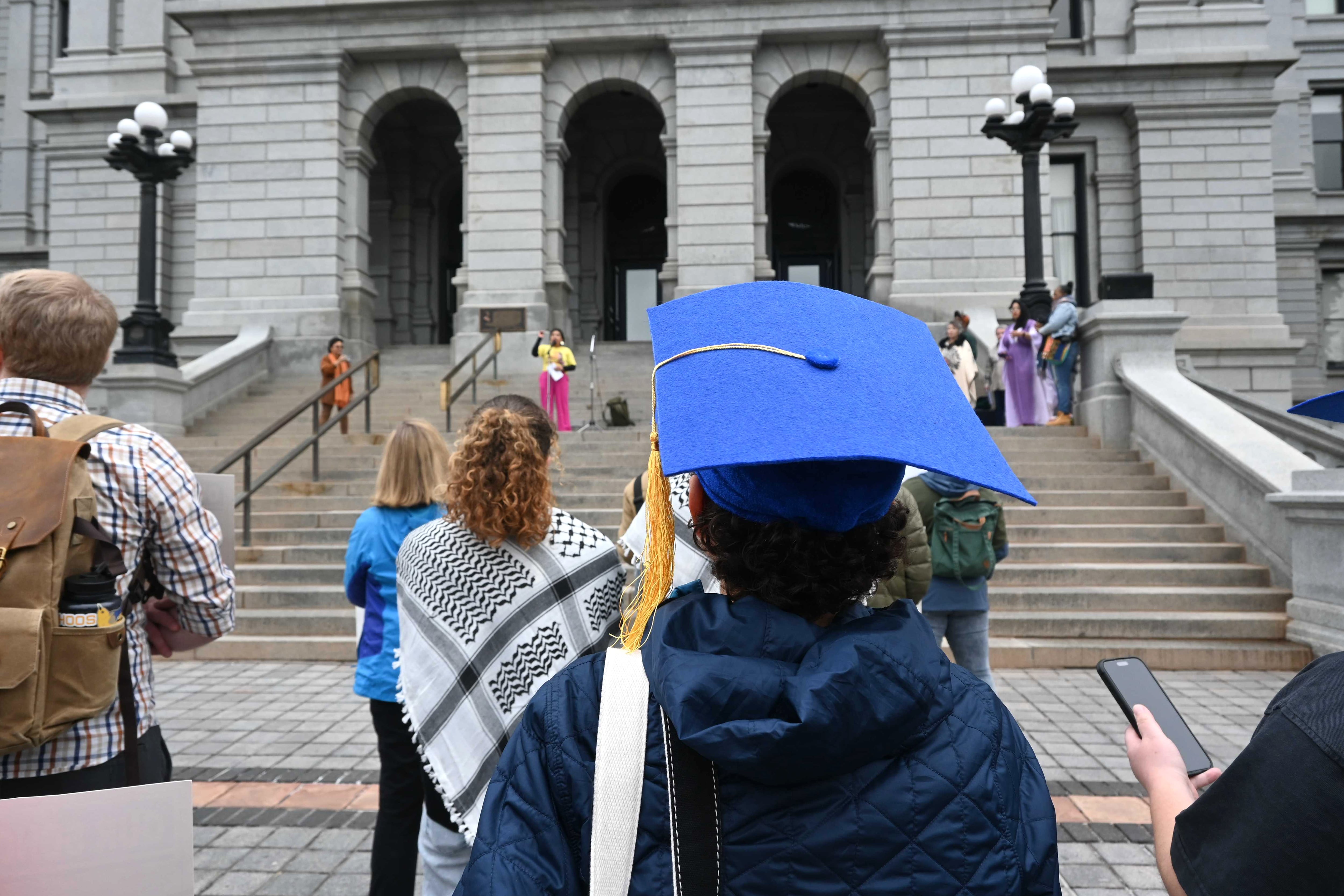 People, including a male in a blue mortarboard, protest on the steps of the Colorado Capitol.