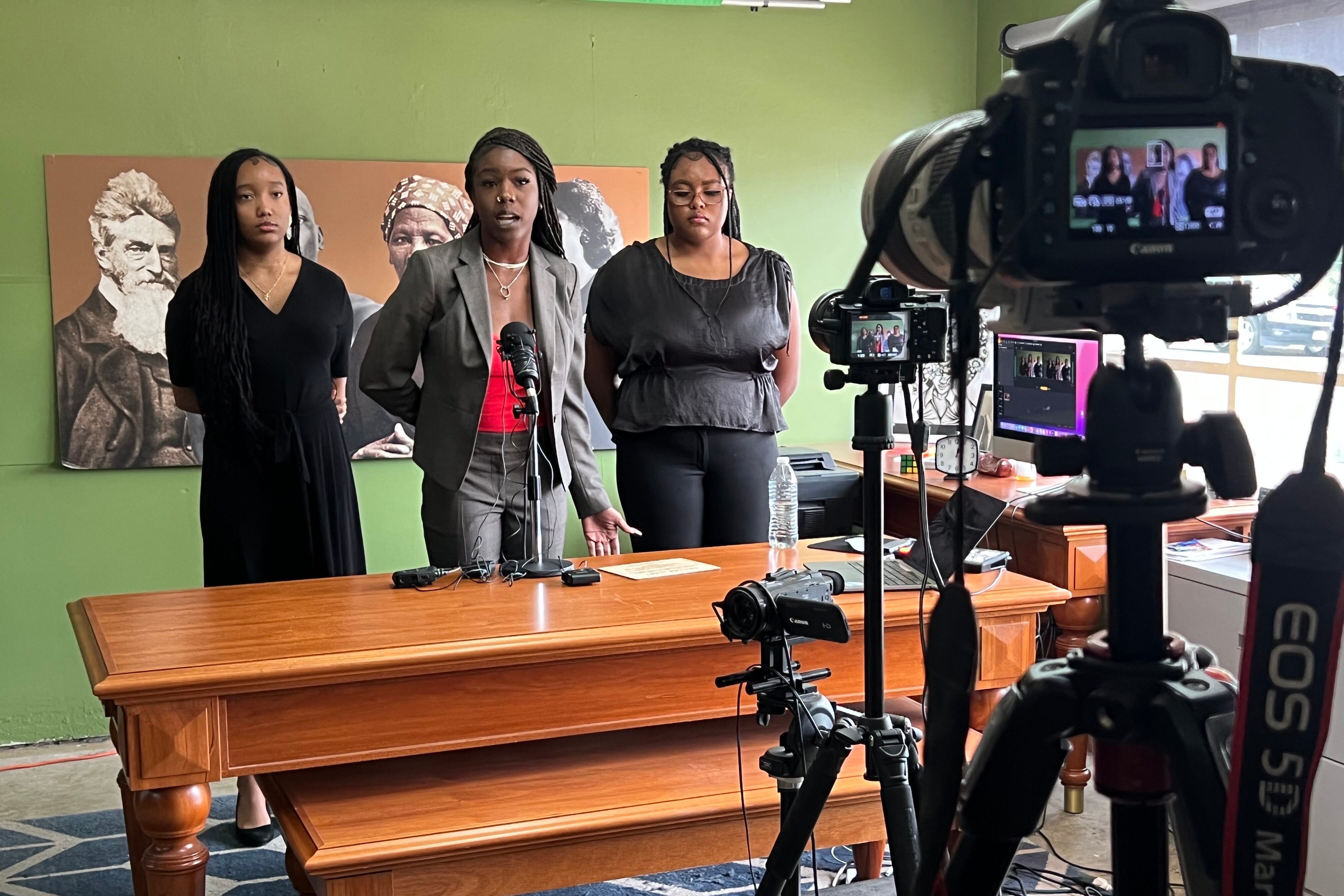 Three young women stand in front of microphones at a press conference.