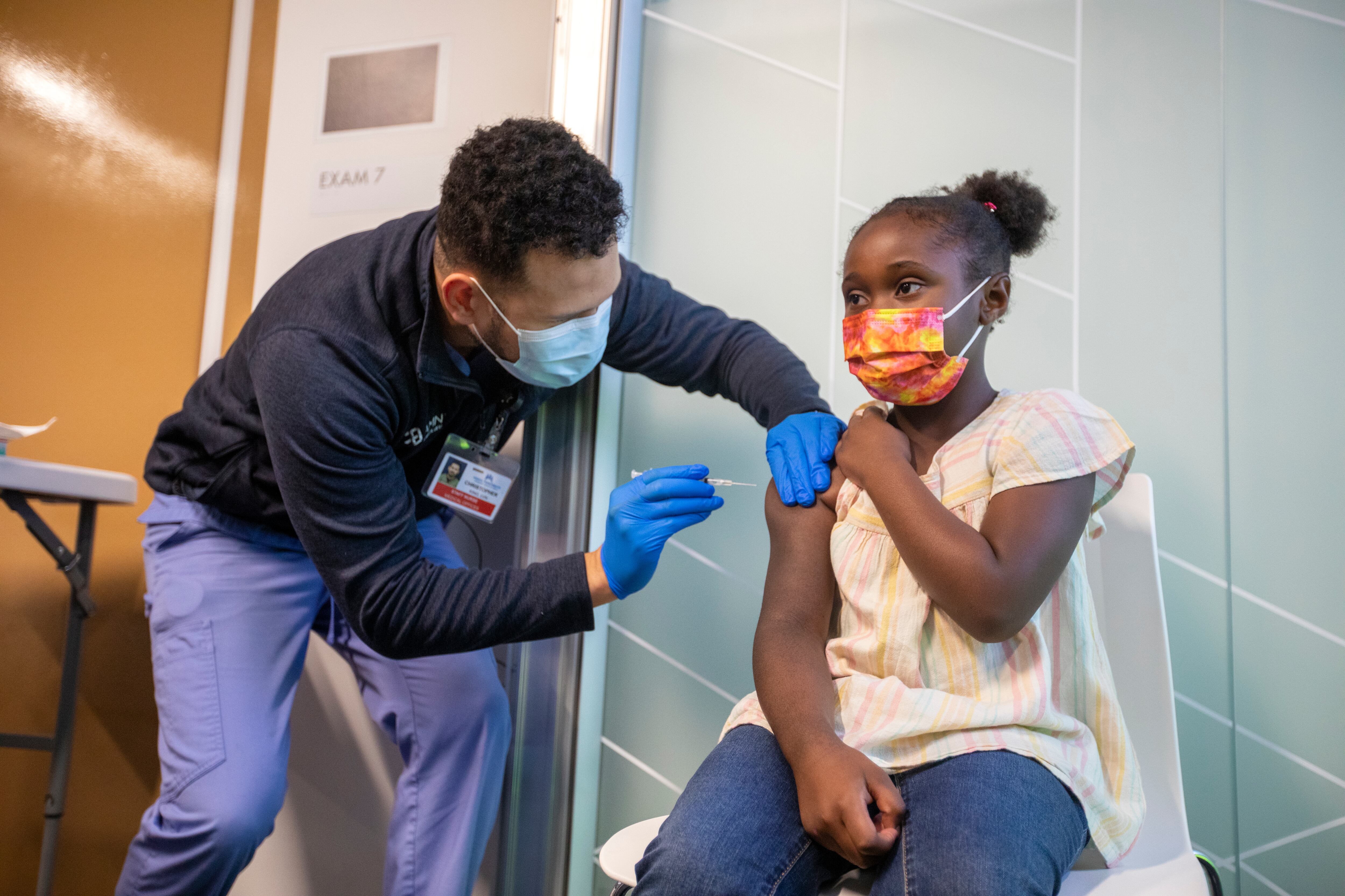 A male health care professional administers a COVID vaccine to a young girl wearing a yellow blouse and blue jeans.