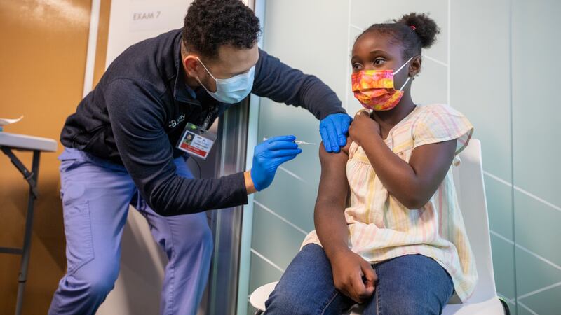 A male health care professional administers a COVID vaccine to a young girl wearing a yellow blouse and blue jeans.