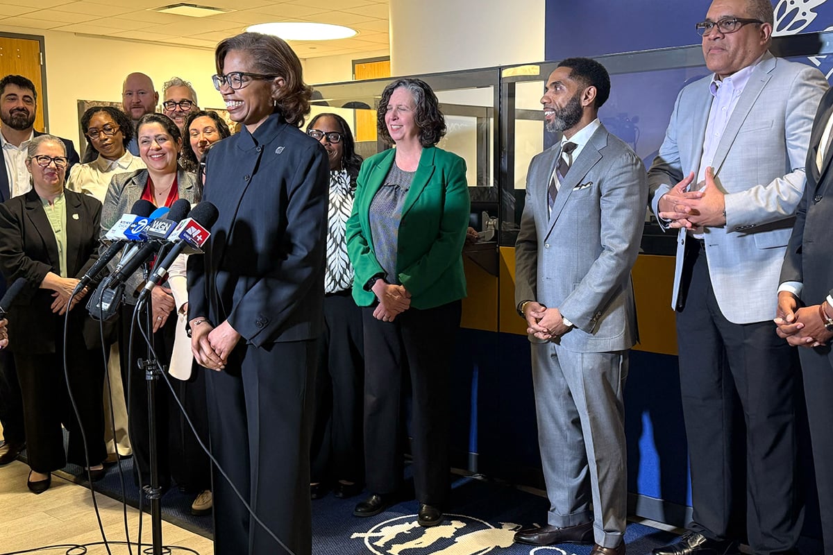 A photograph of a Black woman in a suit standing behind a row of microphones with a row of adults standing behind her.