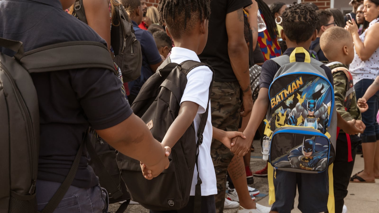 A line of students wearing backpacks hold hands while they walk toward the entrance of school in a crowd of people.