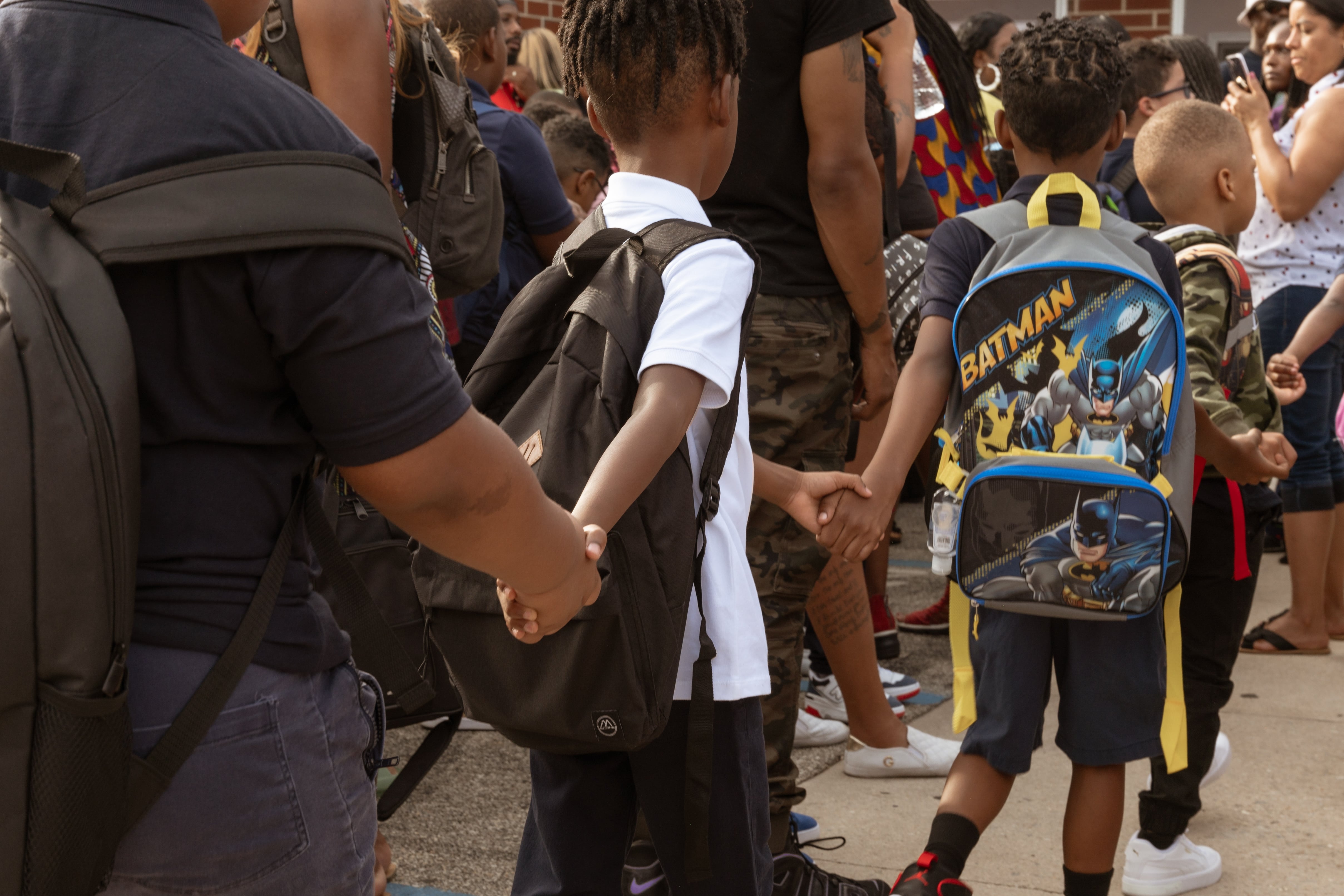 A line of students wearing backpacks hold hands while they walk toward the entrance of school in a crowd of people.