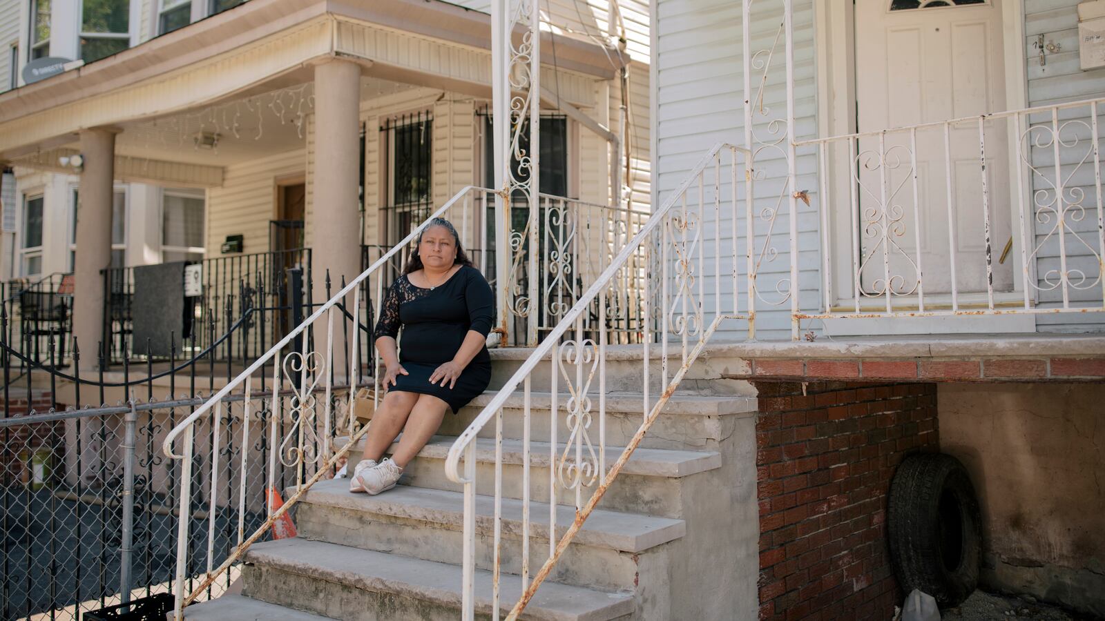 Patricia Coyotecatl, wearing a black dress and white sneakers, sits on the concrete front steps of her home in Newark, which is framed by a white metal fence on either side.