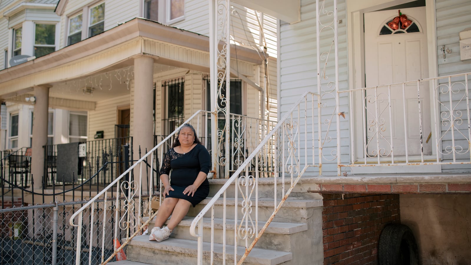 Patricia Coyotecatl, wearing a black dress and white sneakers, sits on the concrete front steps of her home in Newark, which is framed by a white metal fence on either side.