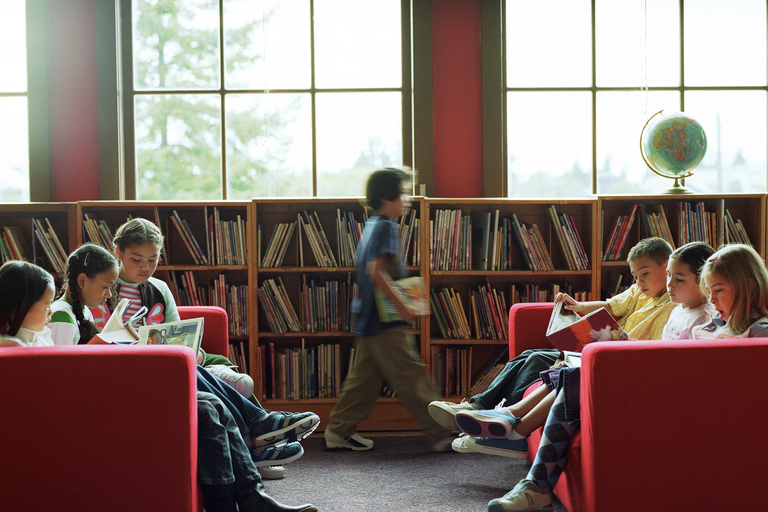 A photograph of six elementary school students, three on each side sitting on two red couches facing each other in a library while one student walks by in the middle.