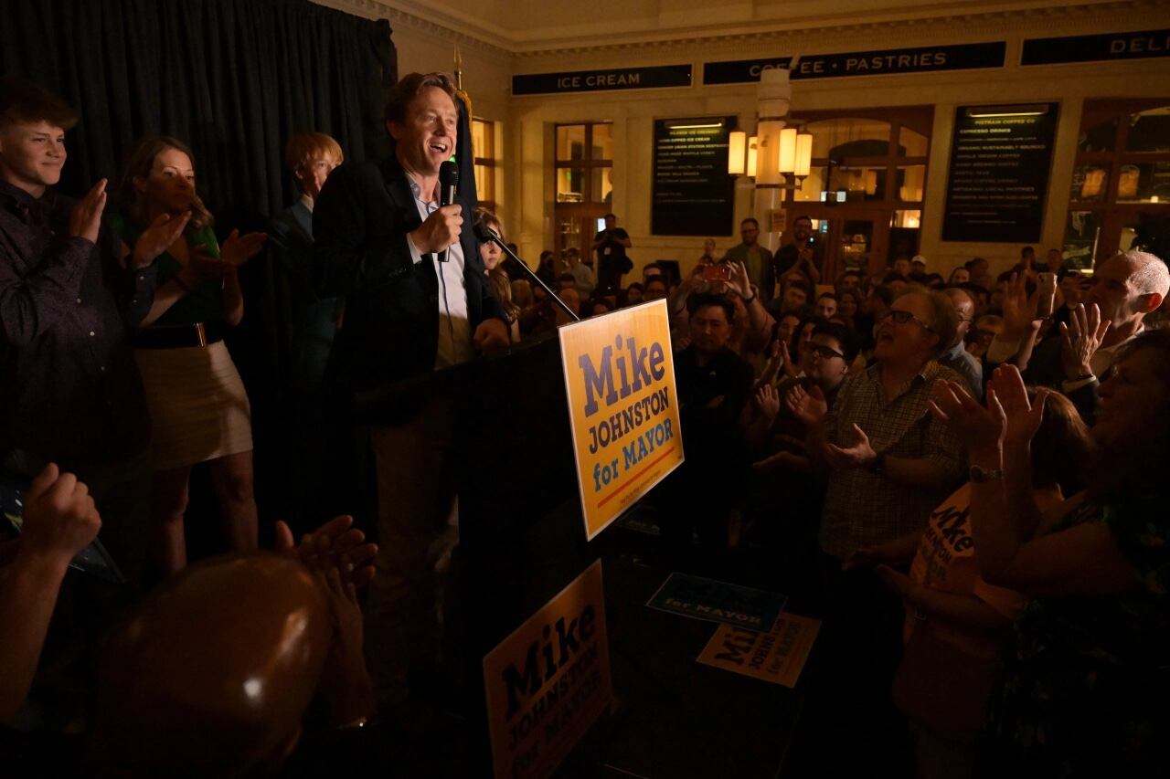 A man, Mike Johnston, stands at a darkened room from a podium with a “Mike Johnston for mayor” sign on front of it and addresses a crowd of people.