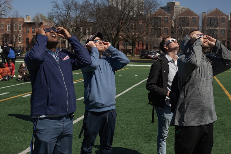 Four adults wearing solar eclipse viewing glasses look up at the sky on a sports field.