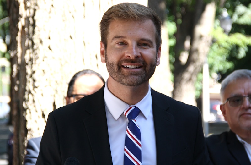 A man in a dark suit jacket and striped tie speaks at a press conference.
