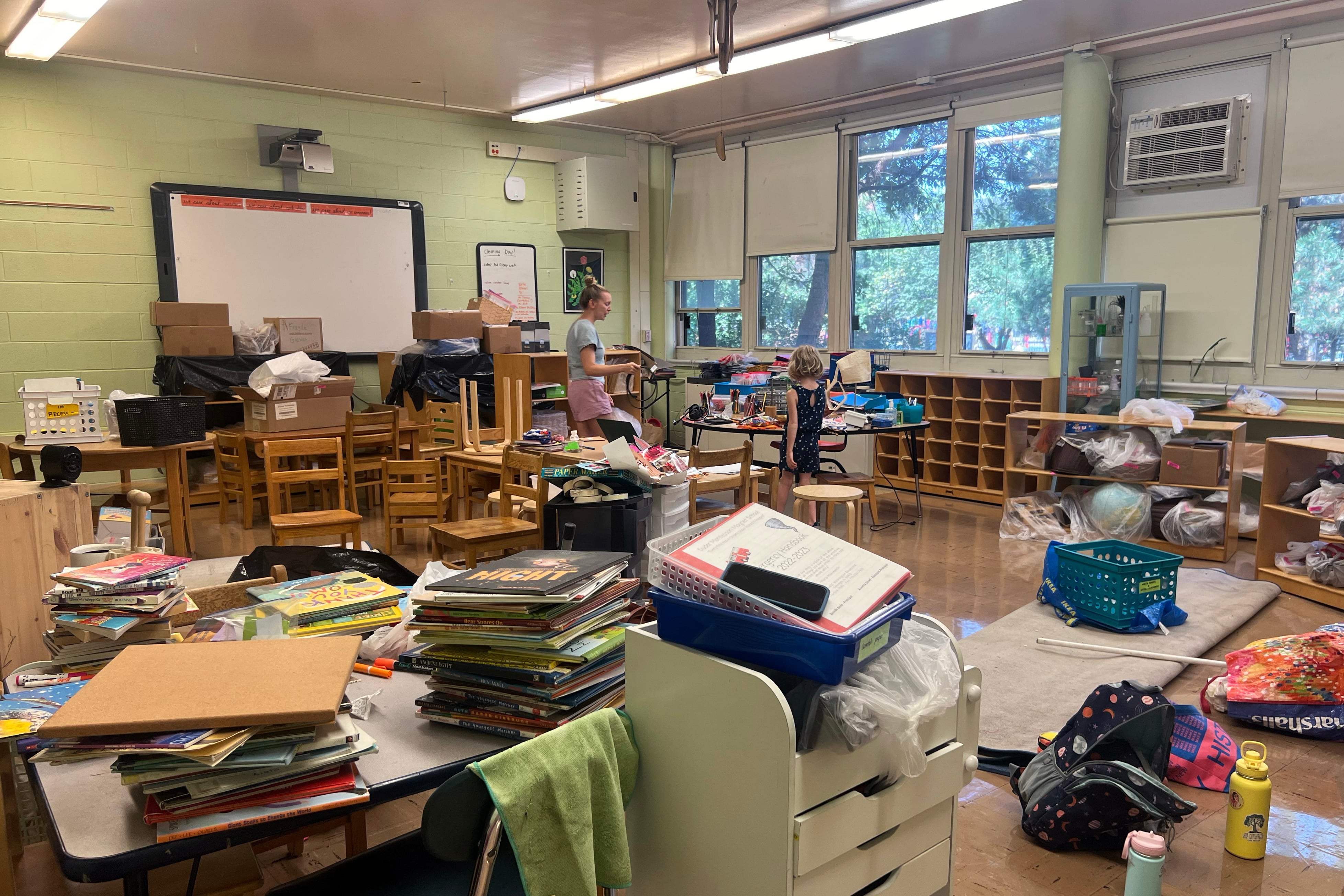 An adult woman and a young child stand in the middle of a cluttered classroom.