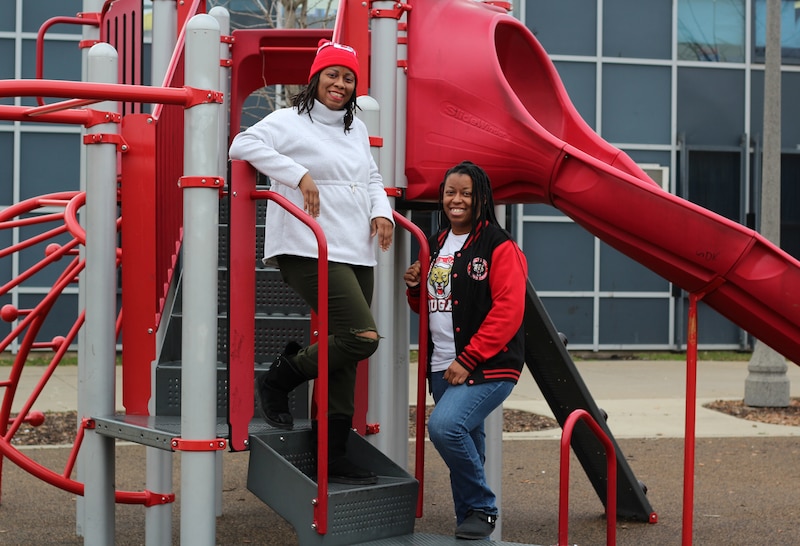 Two women with dark hair stand next to each other outside on a red and white playground outside of a school.