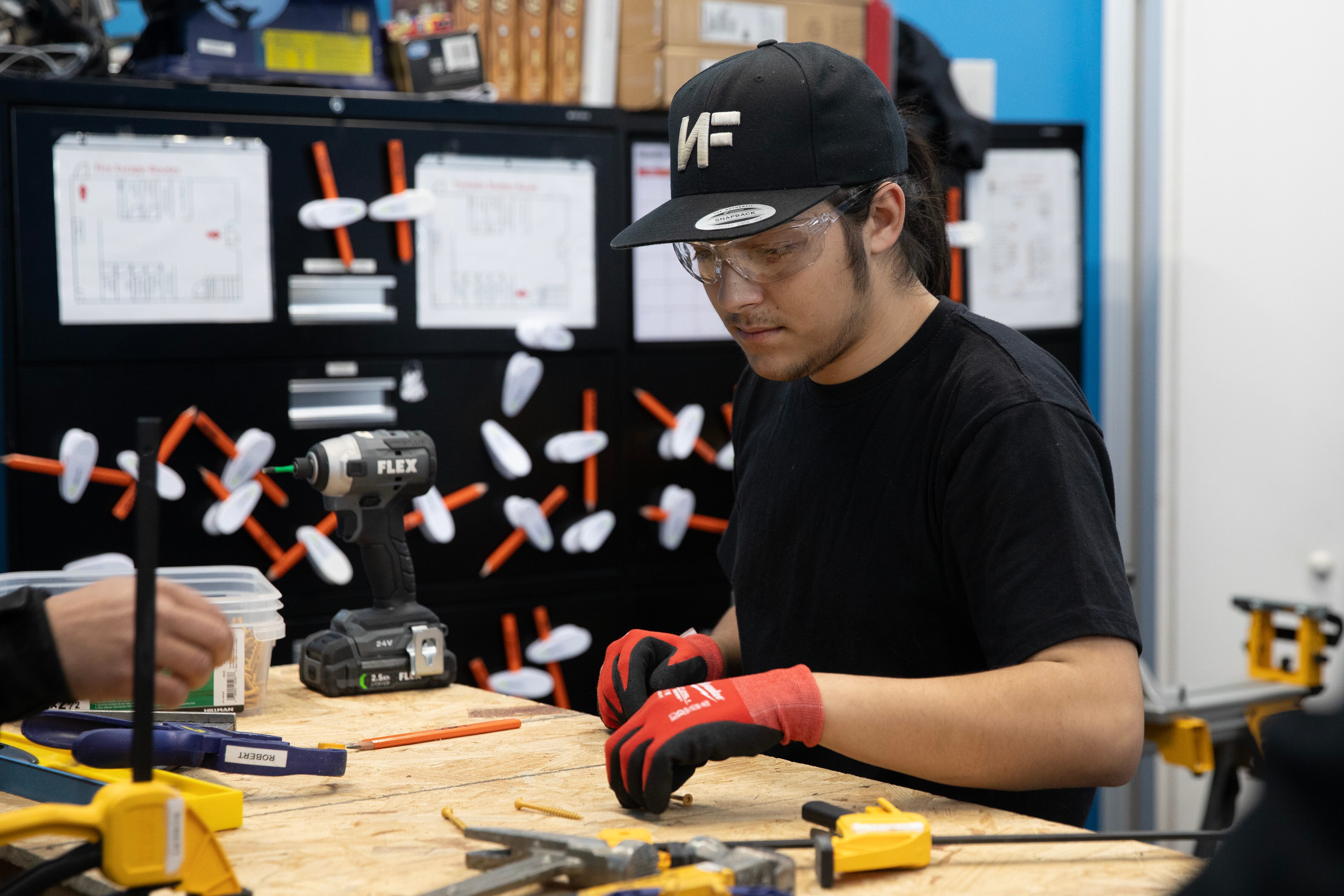 A young adult wearing safety goggles and a baseball hat works during a wood shop class.