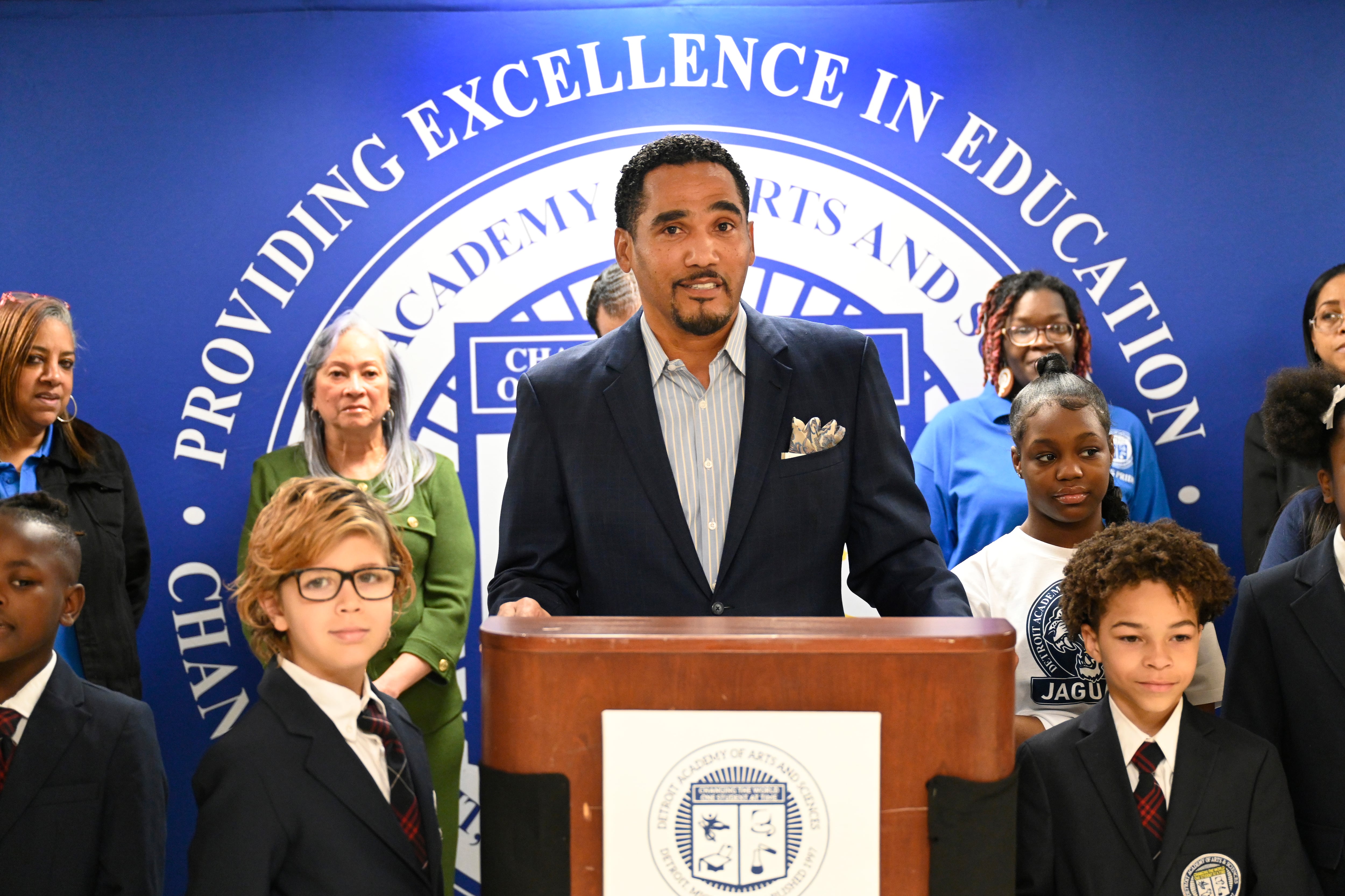 A school leader stands behind a podium, surrounded by students and staff.