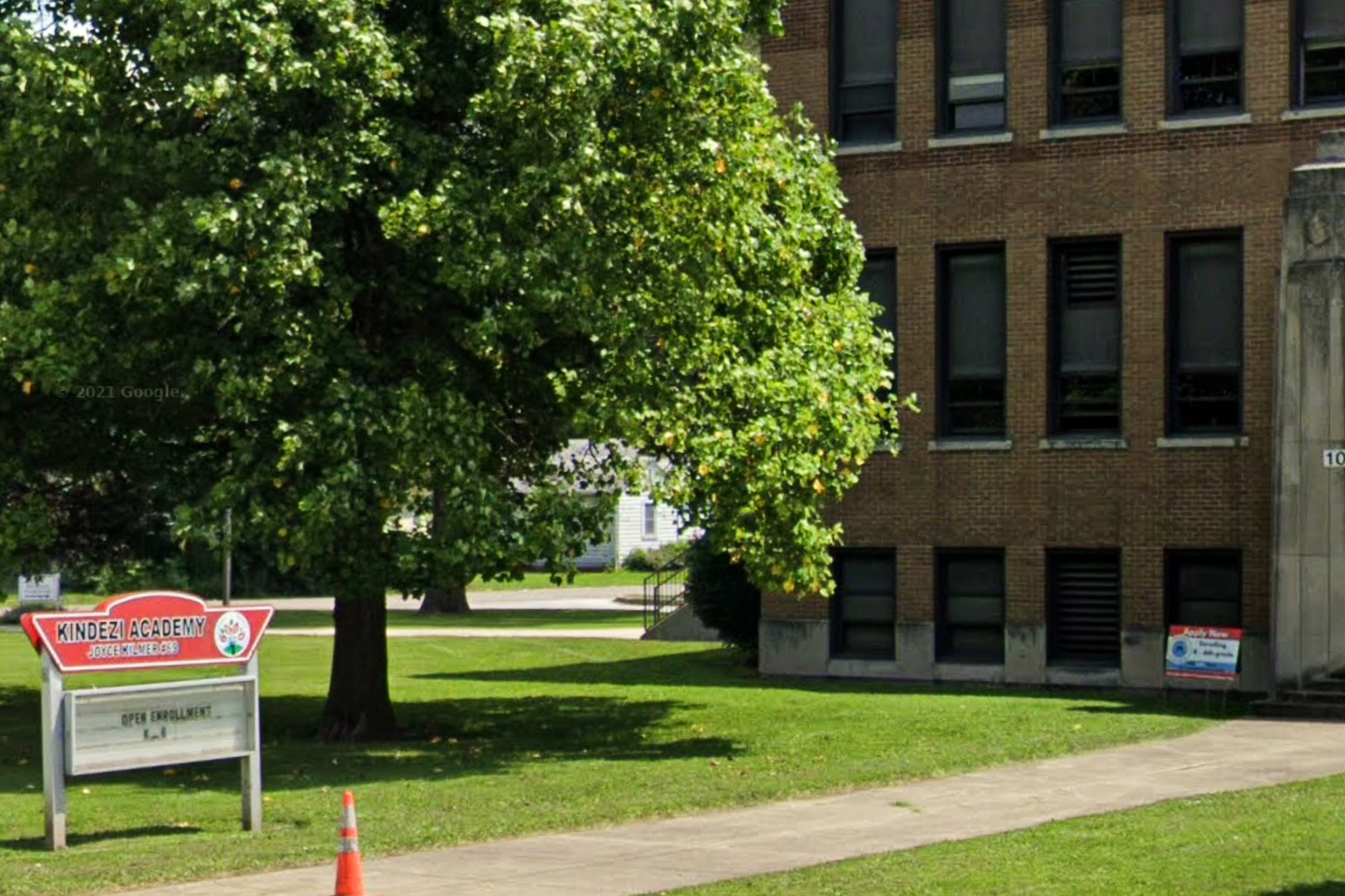 In front of a brick building, a sign for Kindezi Academy at Joyce Kilmer School 69 reads “Open Enrollment K_6.”