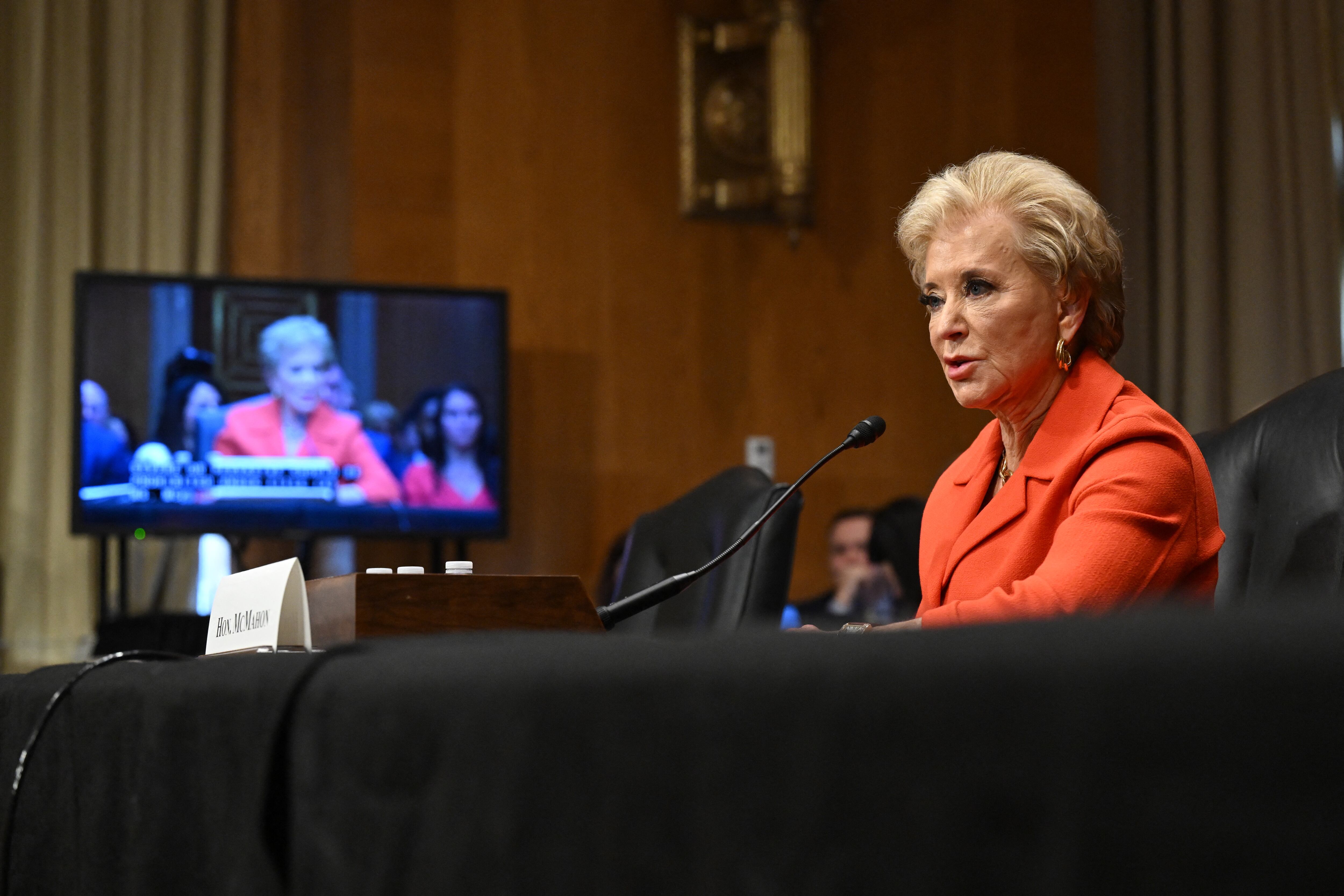 A woman with short white hair and wearing an orange suit sits at a black table talking into a microphone with a TV screen in the background showing her talking.