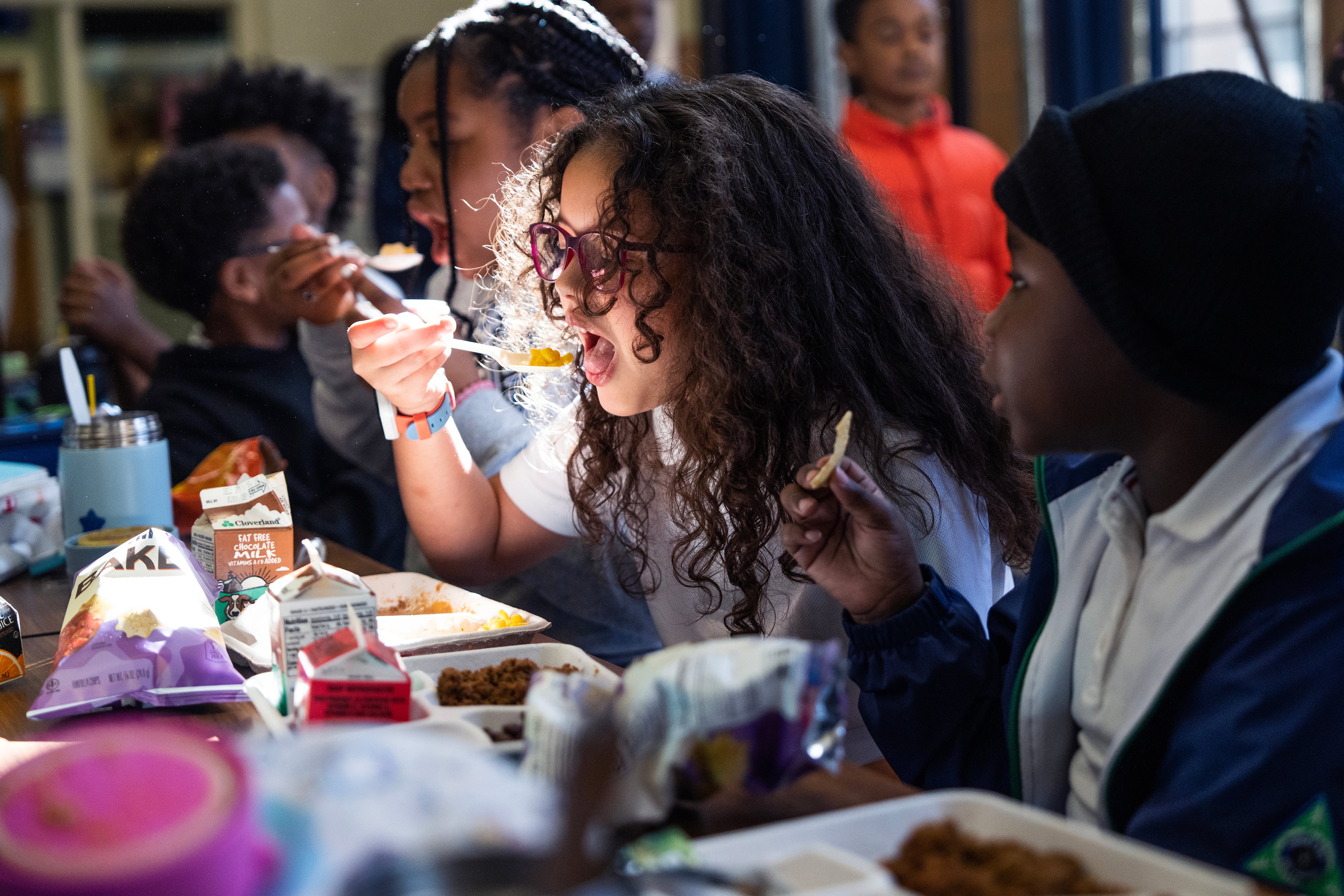A row of elementary students sit at a cafeteria table eating lunch.