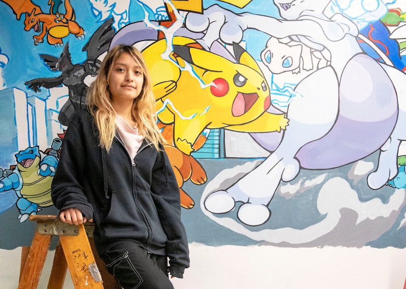 A photograph of a high school student with long blonde hair posing for a portrait on top of a ladder next to a colorful mural.