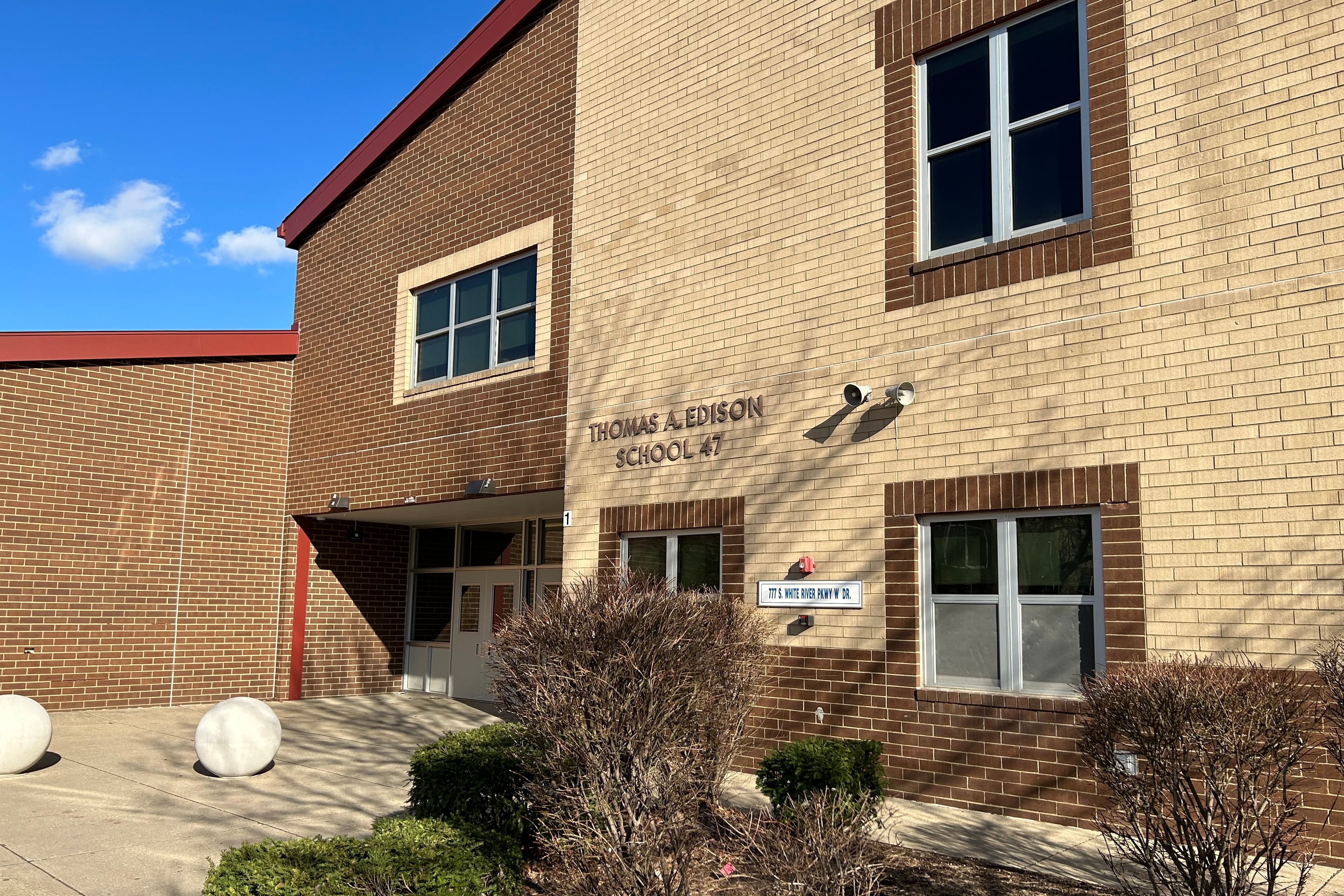 The front of a tan brick building housing Edison School of the Arts rises behind some shrubbery and a patio, against a blue sky.