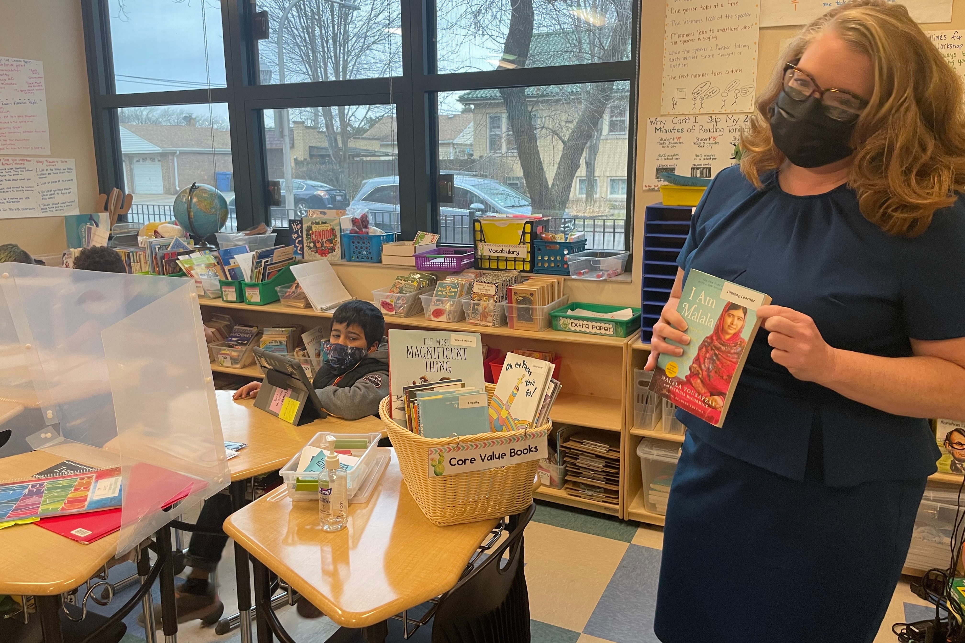 A woman, wearing a blue dress, glasses, and protective mask, hold up the book I Am Malala in an elementary school classroom as students sit at their desks in the background.