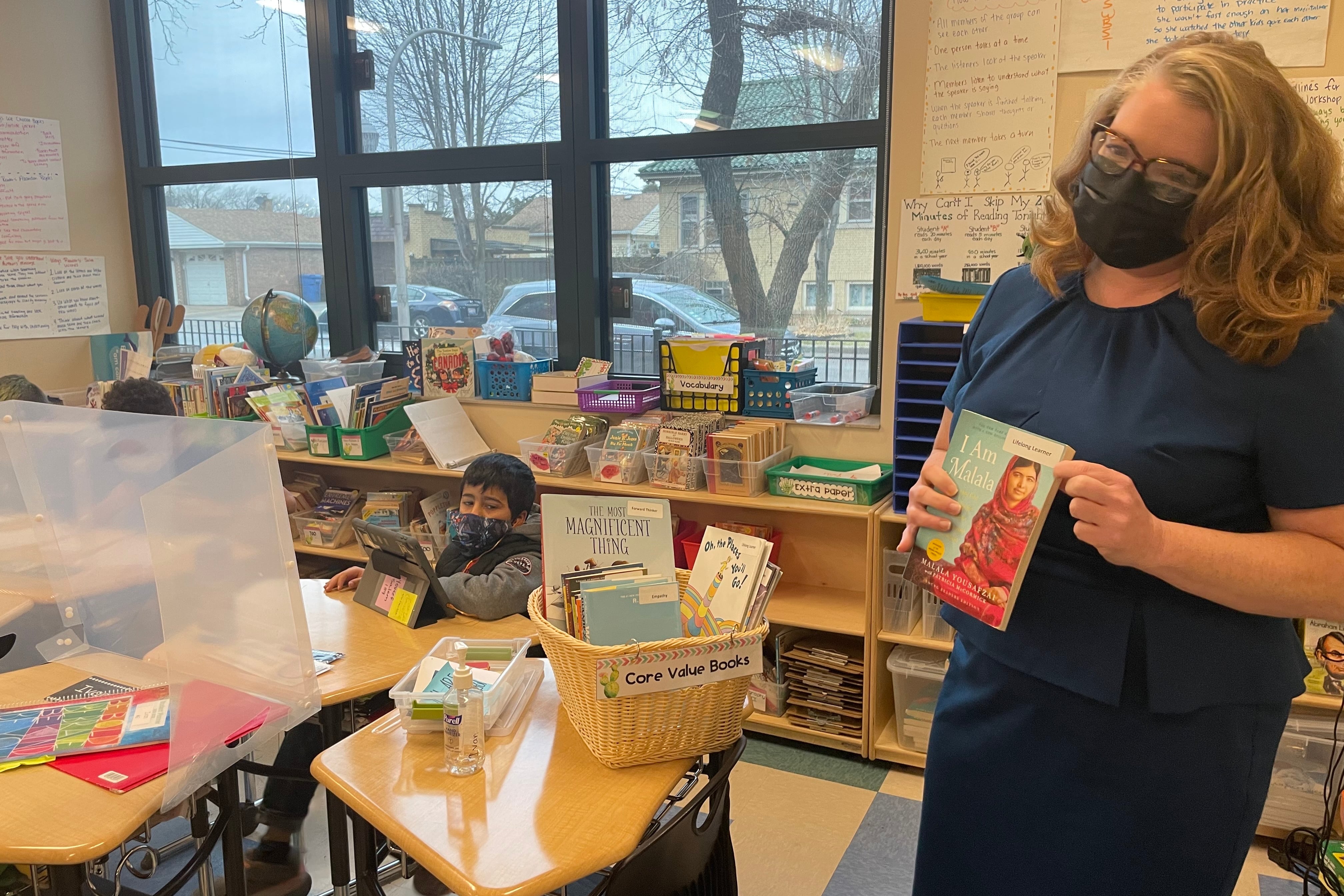 A woman, wearing a blue dress, glasses, and protective mask, hold up the book I Am Malala in an elementary school classroom as students sit at their desks in the background.