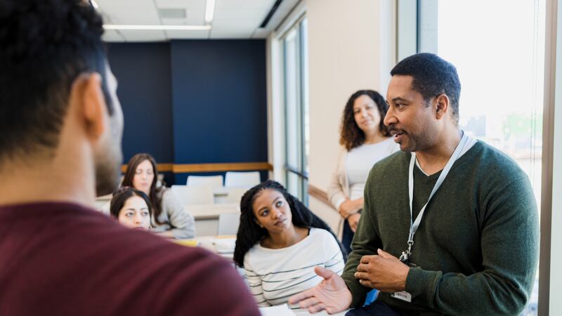 A man in a green shirt and white lanyard talks to a group of people in a brightly lit room.