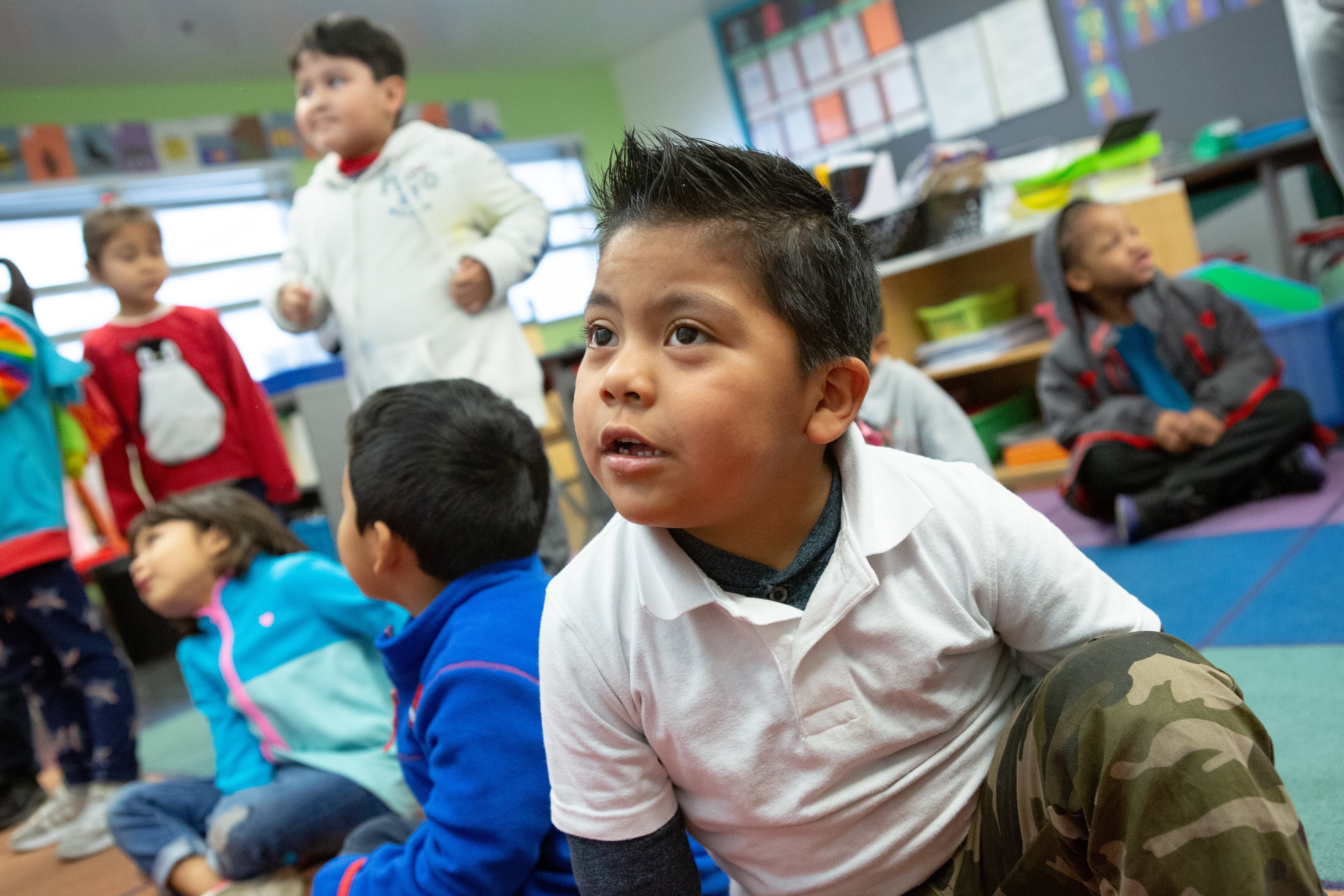 A young boy in a white shirt is sitting on the rug in a classroom with other children.