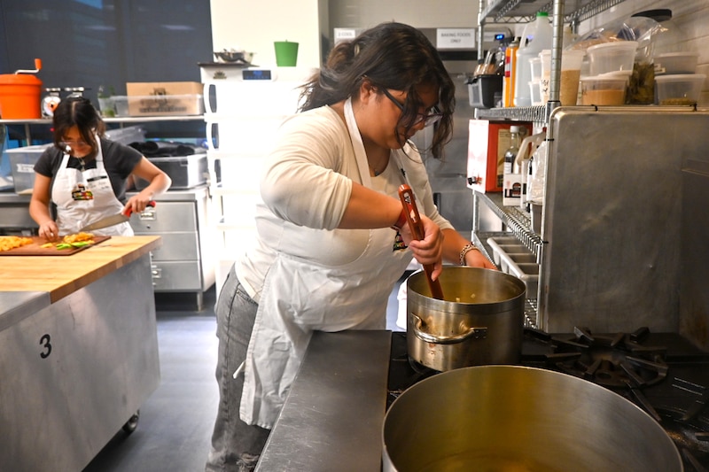 A woman with glasses and wearing a white apron cooks at a stove.