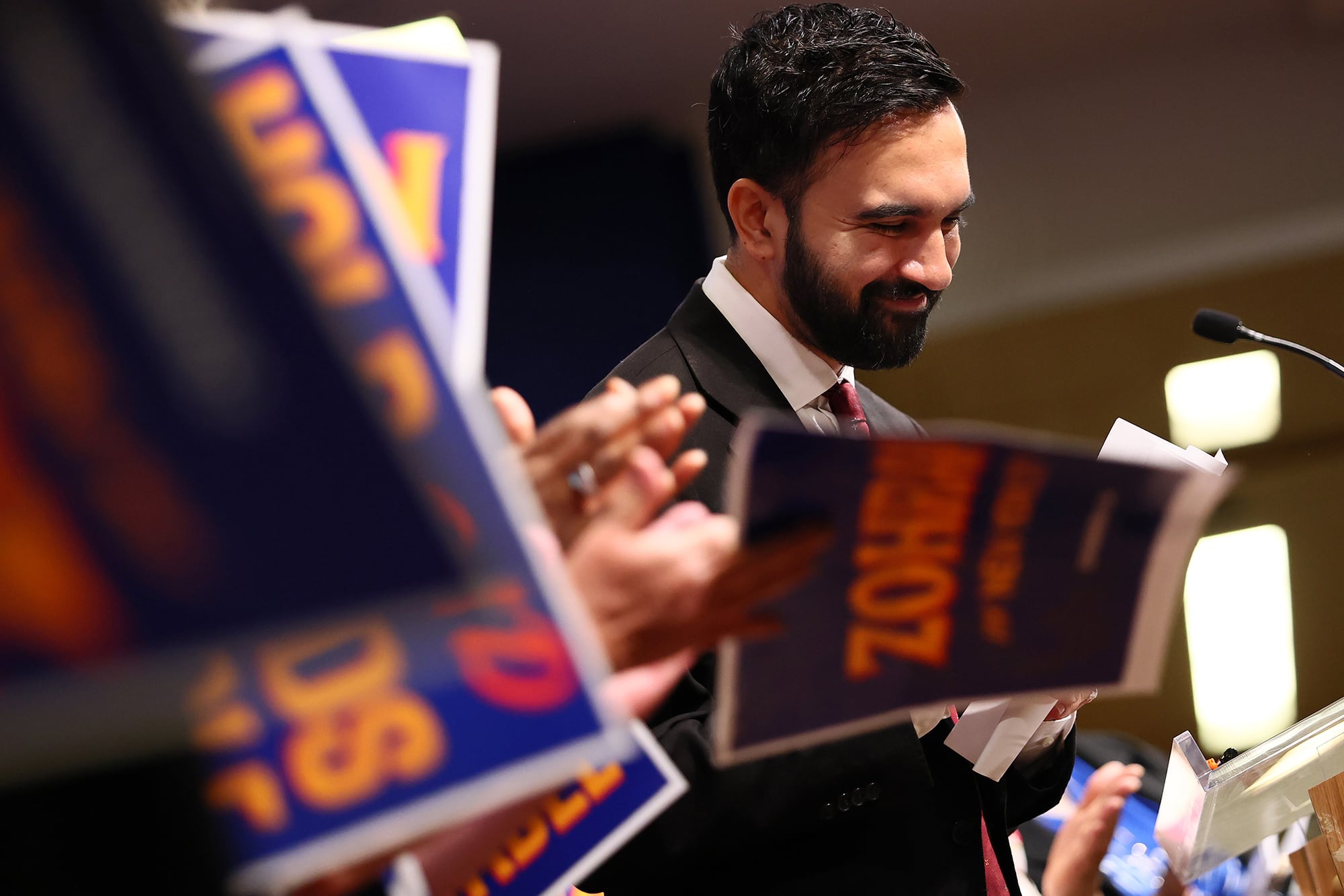 A photograph of a man in a suit behind a row of political signs and a microphone.