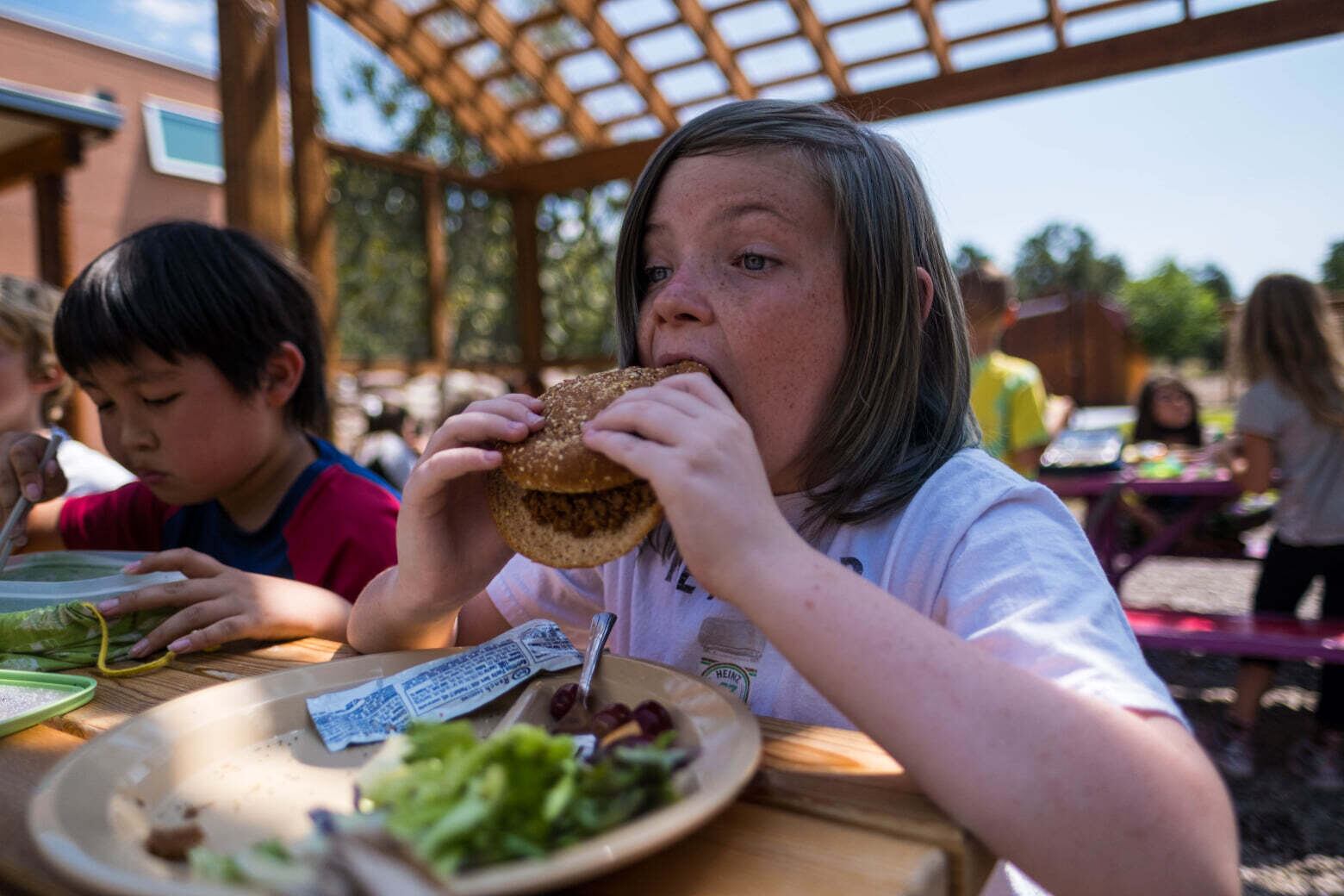 A student at Crestone Charter School eats a hamburger at an outdoor lunch table, as other students sit around them.