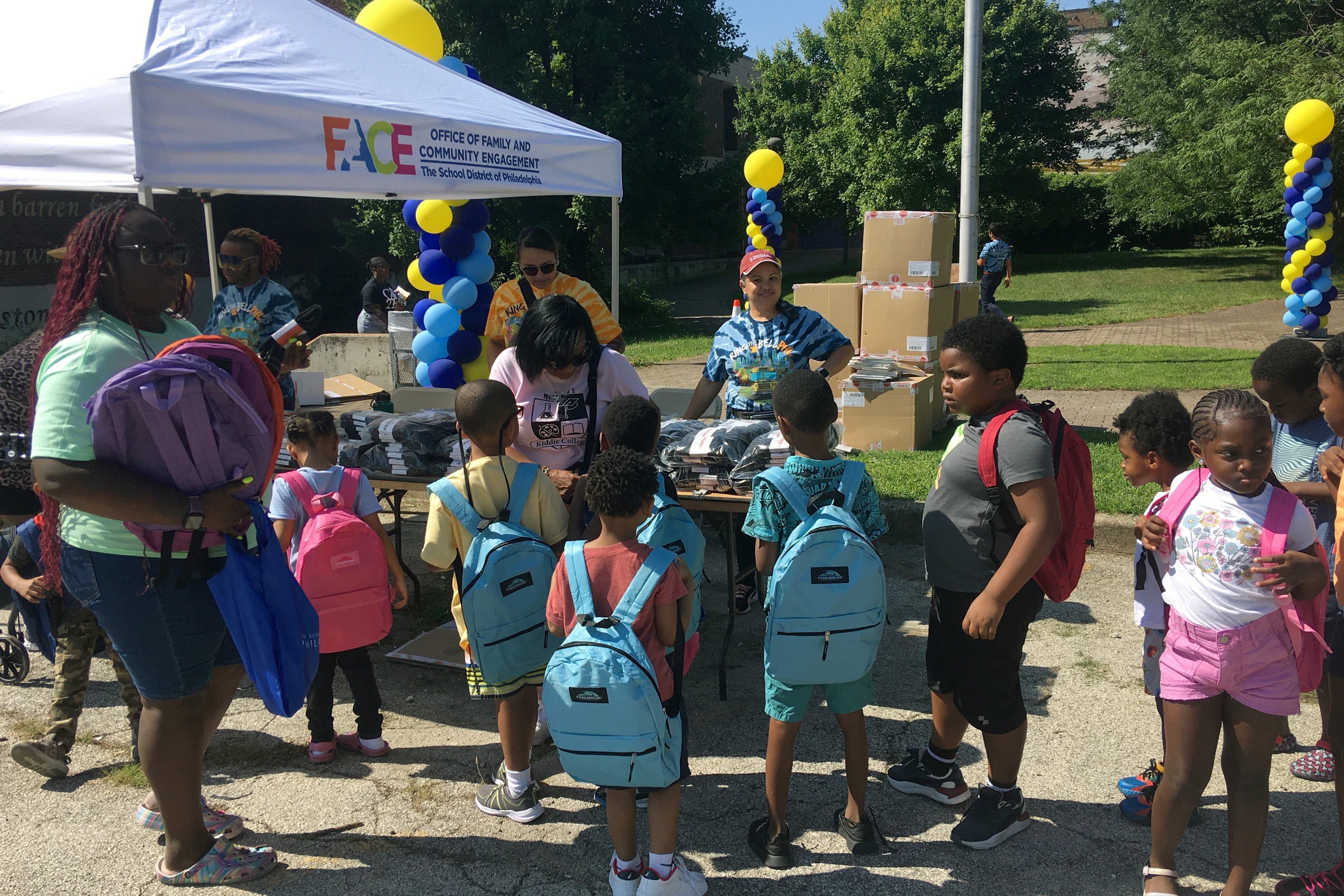 A group of children in large, colorful backpacks stand with their teacher.